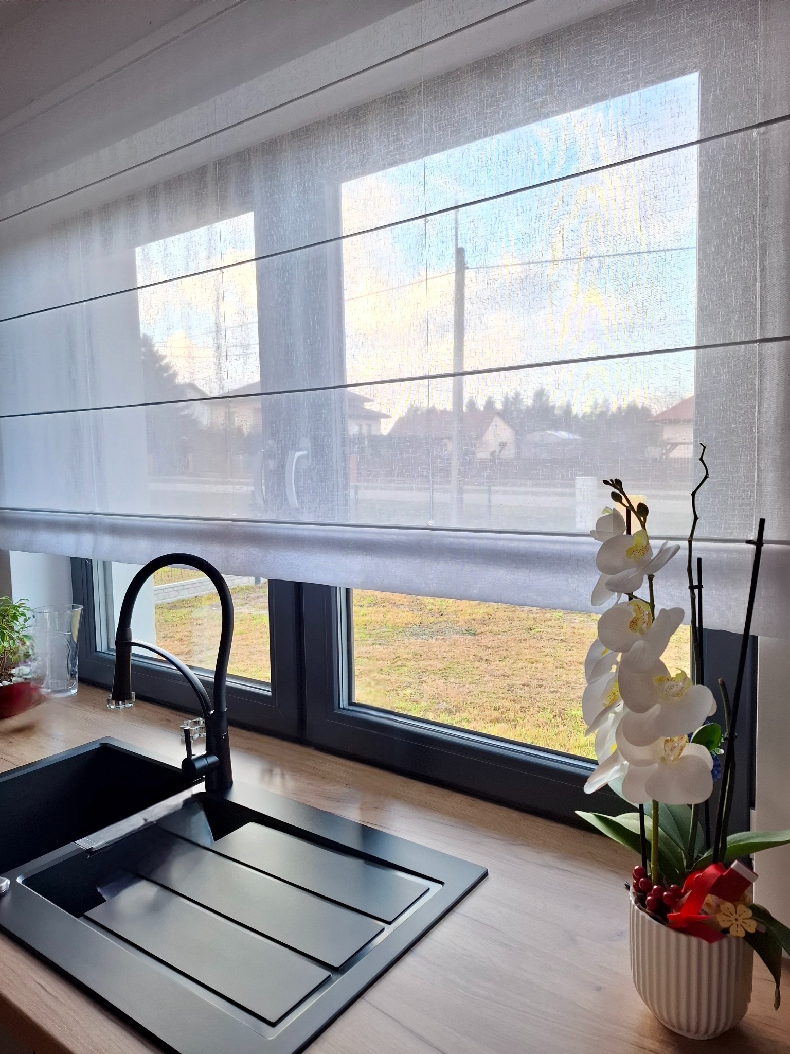 Jasna, zwiewna roleta rzymska w kuchni z czarnym zlewem i kwiatami. Widok z okna na zewnątrz. Delikatne światło dzienne.