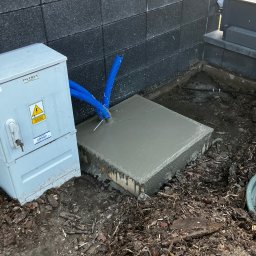 Techniki i technologie komunikacyjne Mariusz Czech - Zabetonowany fundament pod szafkę elektryczną z wystającymi niebieskimi rurami ochronnymi, obok szara szafka elektryczna z ostrzeżeniem o napięciu. Prace instalacyjne.