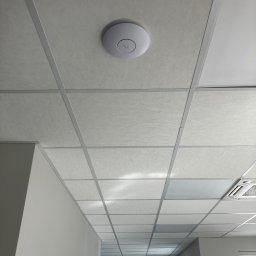 Techniki i technologie komunikacyjne Mariusz Czech - Biały punkt dostępowy Wi-Fi zamontowany na kasetonowym suficie podwieszanym w biurze. Widoczna kratownica sufitowa i fragment klimatyzacji.