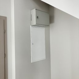 Techniki i technologie komunikacyjne Mariusz Czech - Szara skrzynka z kluczem i biała szafka na ścianie w jasnym pomieszczeniu, prawdopodobnie klatka schodowa. Widoczne elementy instalacji.