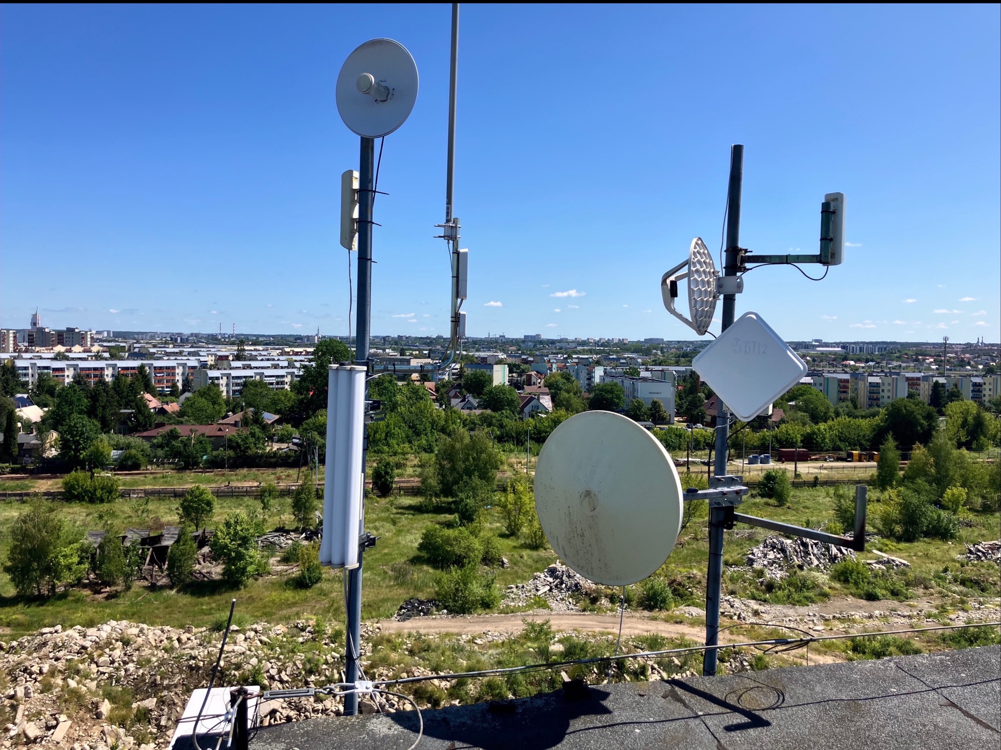 Anteny telekomunikacyjne na dachu budynku w Porosłach, widok na miasto w tle. Różne typy anten, w tym paraboliczne i sektorowe, zamontowane na metalowych masztach.