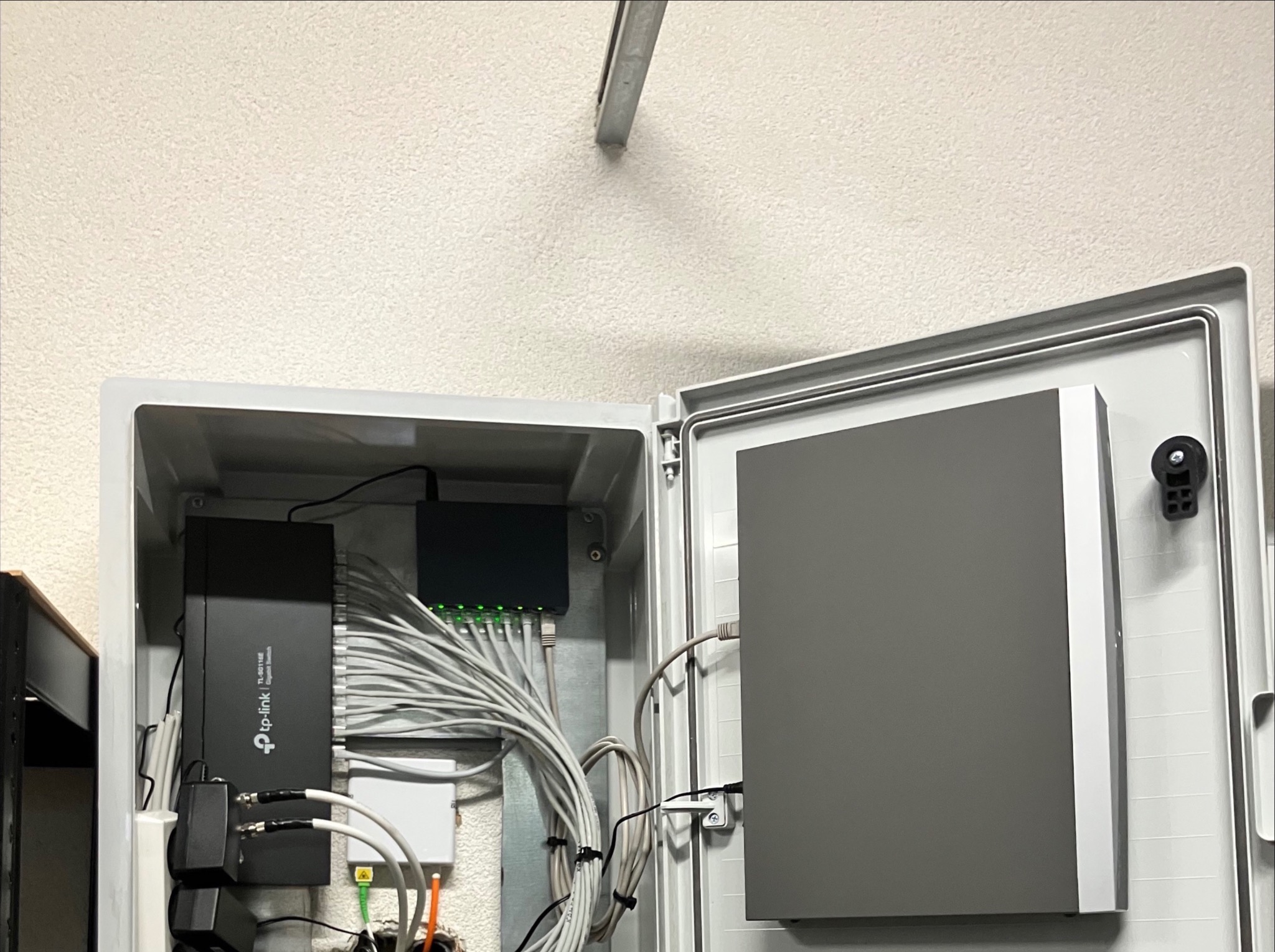 Otwarta skrzynka z urządzeniami sieciowymi TP-Link i okablowaniem, zamontowana na ścianie. Widoczne porty Ethernet i zasilacze. Oświetlenie sufitowe w tle.
