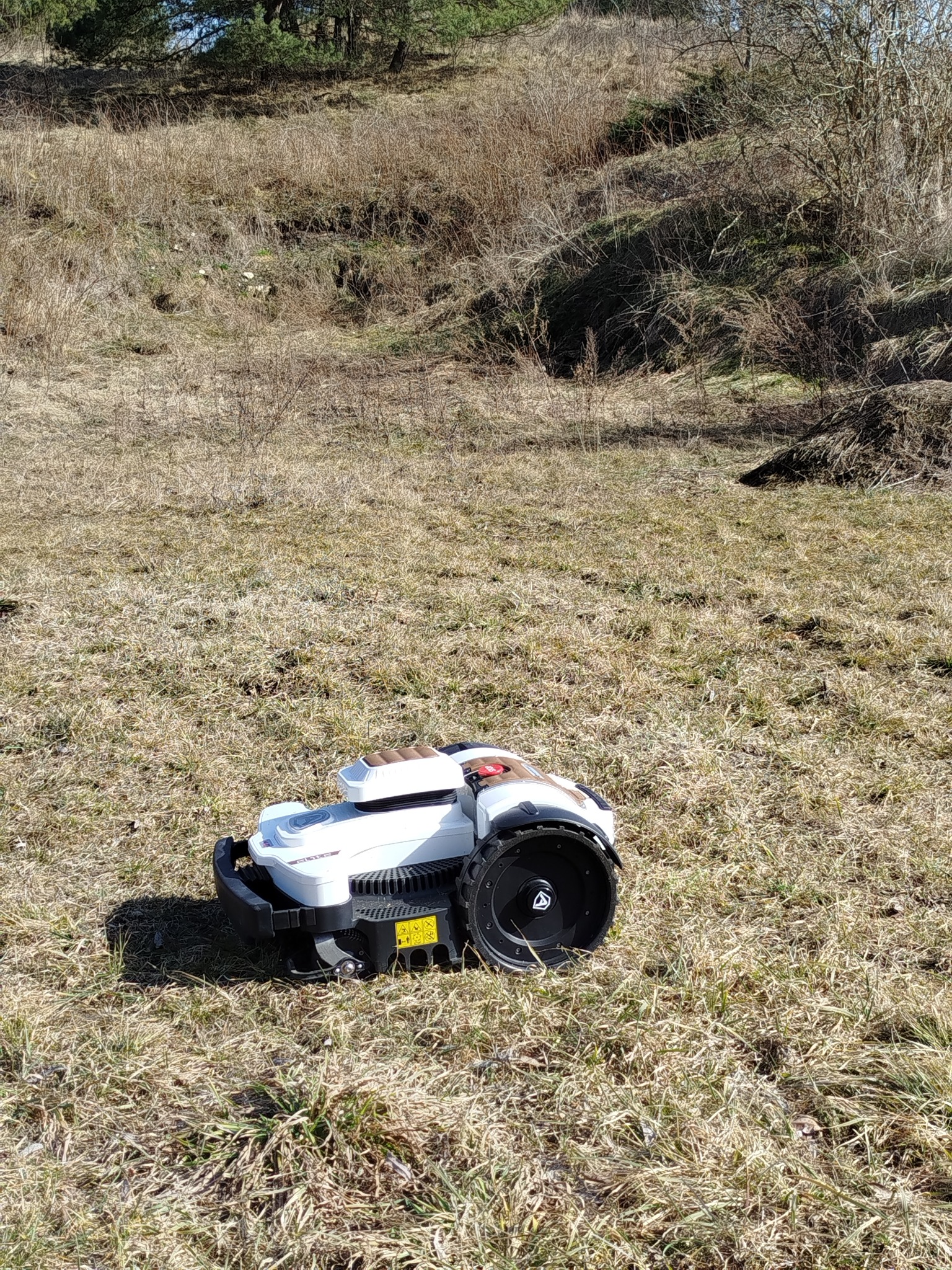 Biały robot koszący na trawniku z suchą trawą, na tle wzgórza i drzew. Widoczne duże koła i elementy ostrzegawcze. Słoneczny dzień.