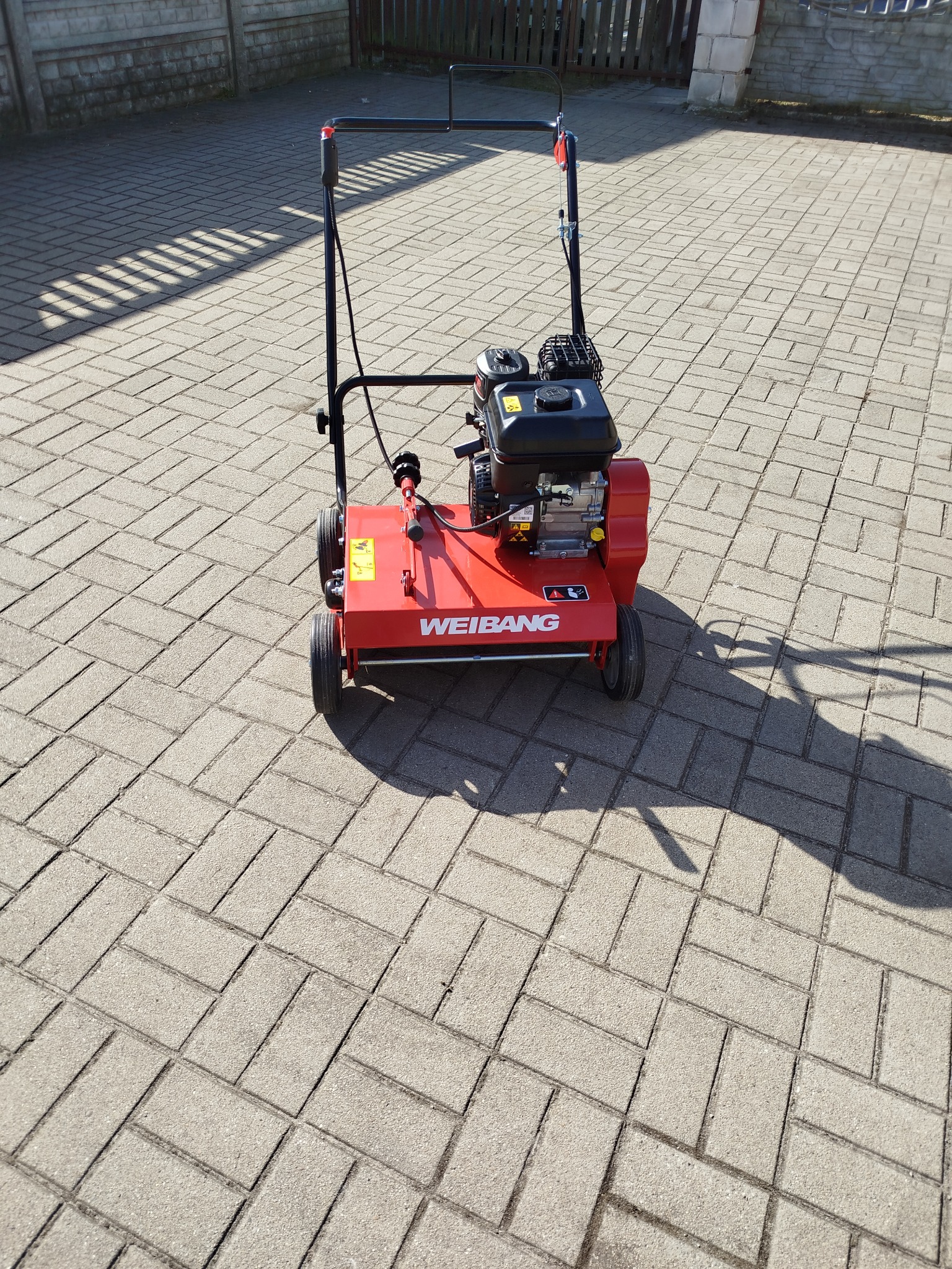 Aerator spalinowy Weibang na kostce brukowej, rzucający cień. Urządzenie w kolorze czerwonym z czarnym silnikiem i uchwytem. Widoczne ślady użytkowania.