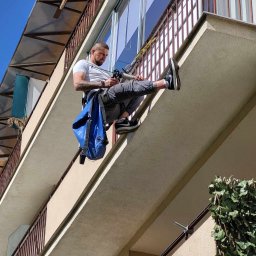 ASSAMAH-VH Vasyl Habiak - Pracownik na linie, zabezpieczony uprzężą, uszczelnia silikonem balkon w budynku wielorodzinnym. Widok z dołu na elewację i barierki.