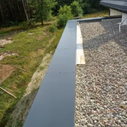 Płaski dach pokryty drobnymi kamieniami, zabezpieczony szarą, metalową obróbką blacharską. Widok z góry na krawędź dachu i otaczającą zieleń.