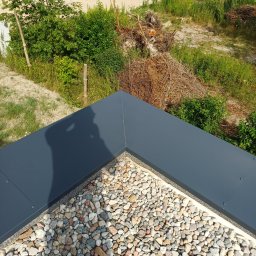 ASSAMAH-VH Vasyl Habiak - Szczegółowe ujęcie dachu pokrytego drobnymi kamieniami, z ciemną, metalową obróbką blacharską w rogu, cień osoby na dachu i zieleń w tle.