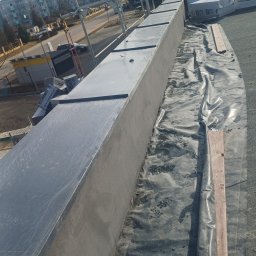 ASSAMAH-VH Vasyl Habiak - Wykończony fragment dachu płaskiego z obróbką blacharską i izolacją, widok z góry, na tle zabudowy miejskiej w słoneczny dzień. Elementy rusztowania w tle.