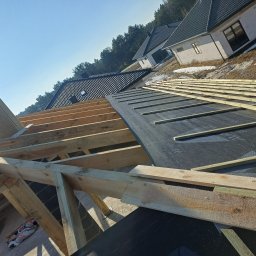 ASSAMAH-VH Vasyl Habiak - Konstrukcja dachu w trakcie budowy: drewniane krokwie, czarna membrana dachowa, widok na domy w tle. Etap prac dekarskich w słoneczny dzień.