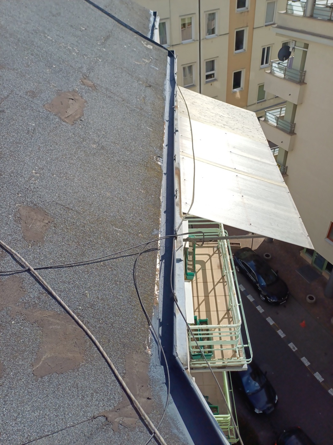 Widok z góry na fragment dachu pokrytego papą, z widoczną krawędzią i budynkami w tle. Uwagę zwraca narożnik dachu z obróbką blacharską.