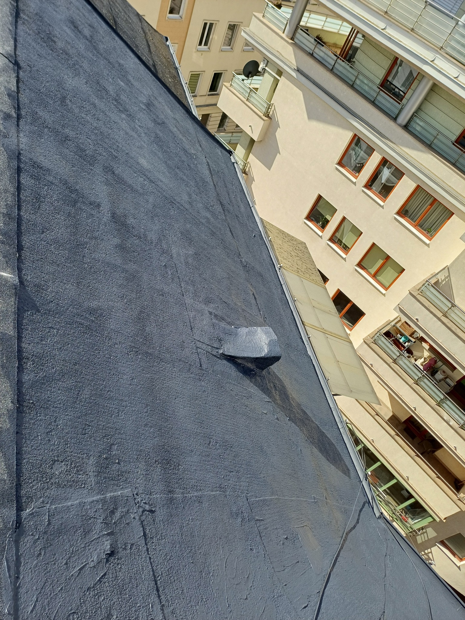 Szary, płaski dach po renowacji, widoczny z góry pod kątem, z budynkami w tle. Na dachu widoczna obróbka blacharska komina.