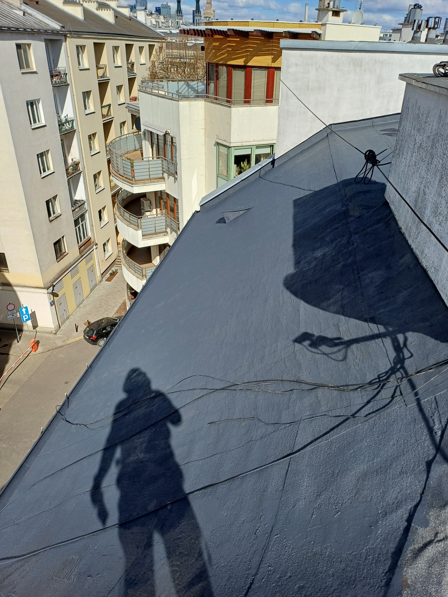 Szary dach z papy widziany z góry, cień osoby i anteny, budynki w tle.  Widok na dach w słoneczny dzień w miejskiej scenerii.
