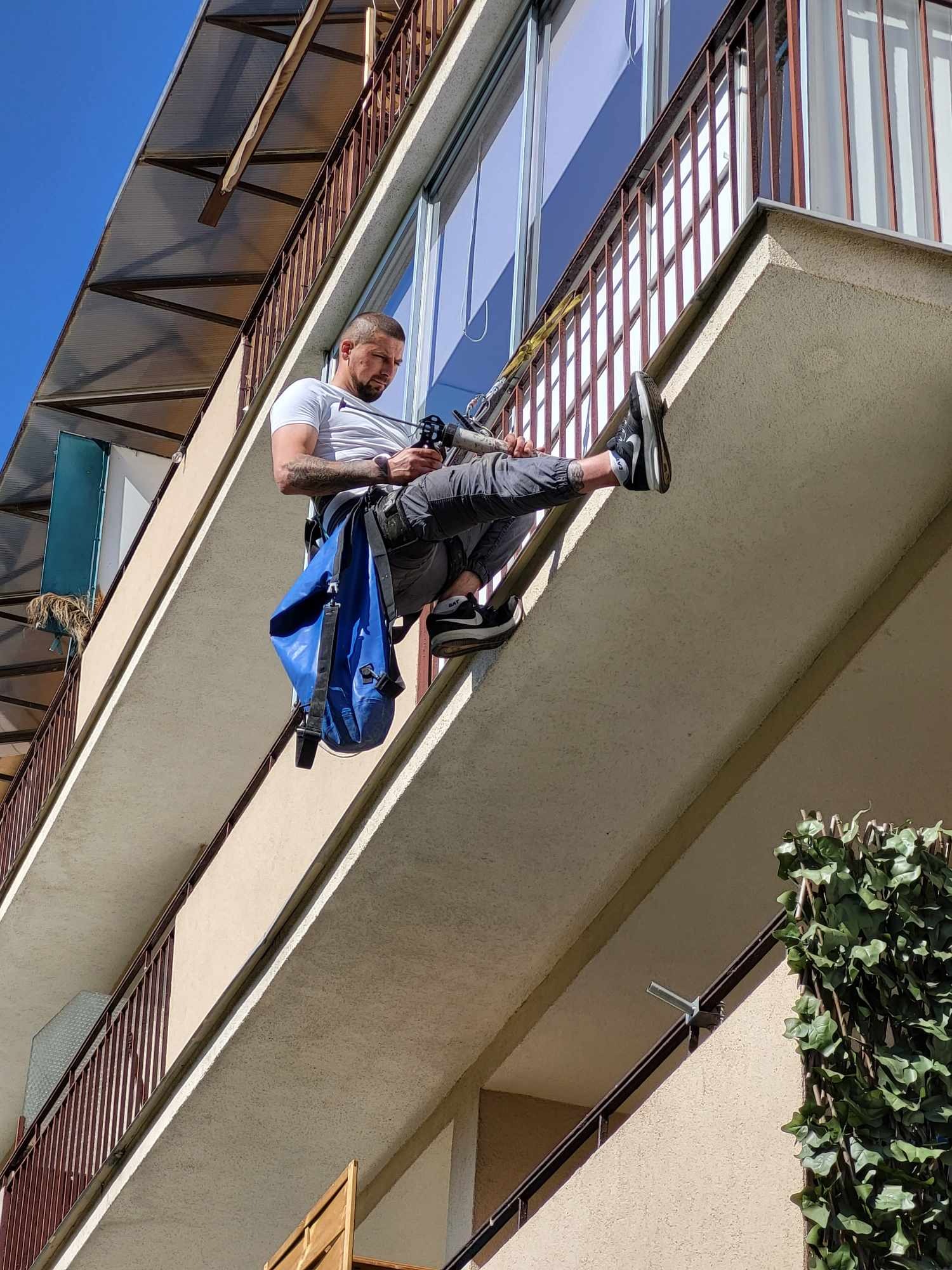 Pracownik na linie, zabezpieczony uprzężą, uszczelnia silikonem balkon w budynku wielorodzinnym. Widok z dołu na elewację i barierki.