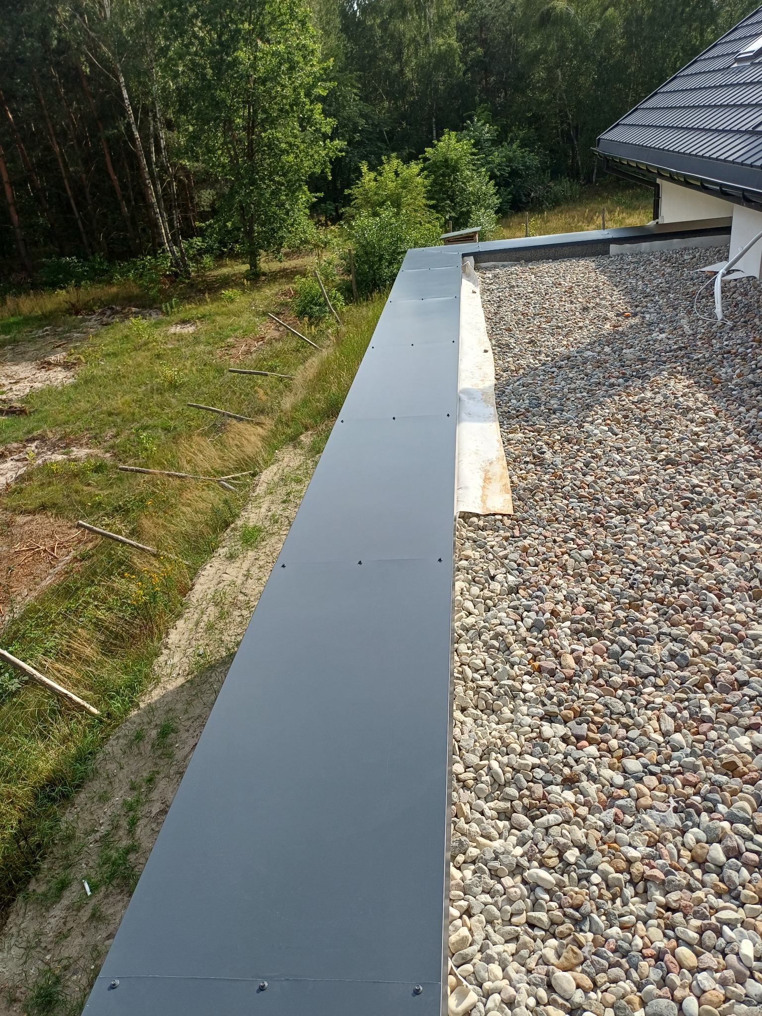 Płaski dach pokryty drobnymi kamieniami, zabezpieczony szarą, metalową obróbką blacharską. Widok z góry na krawędź dachu i otaczającą zieleń.