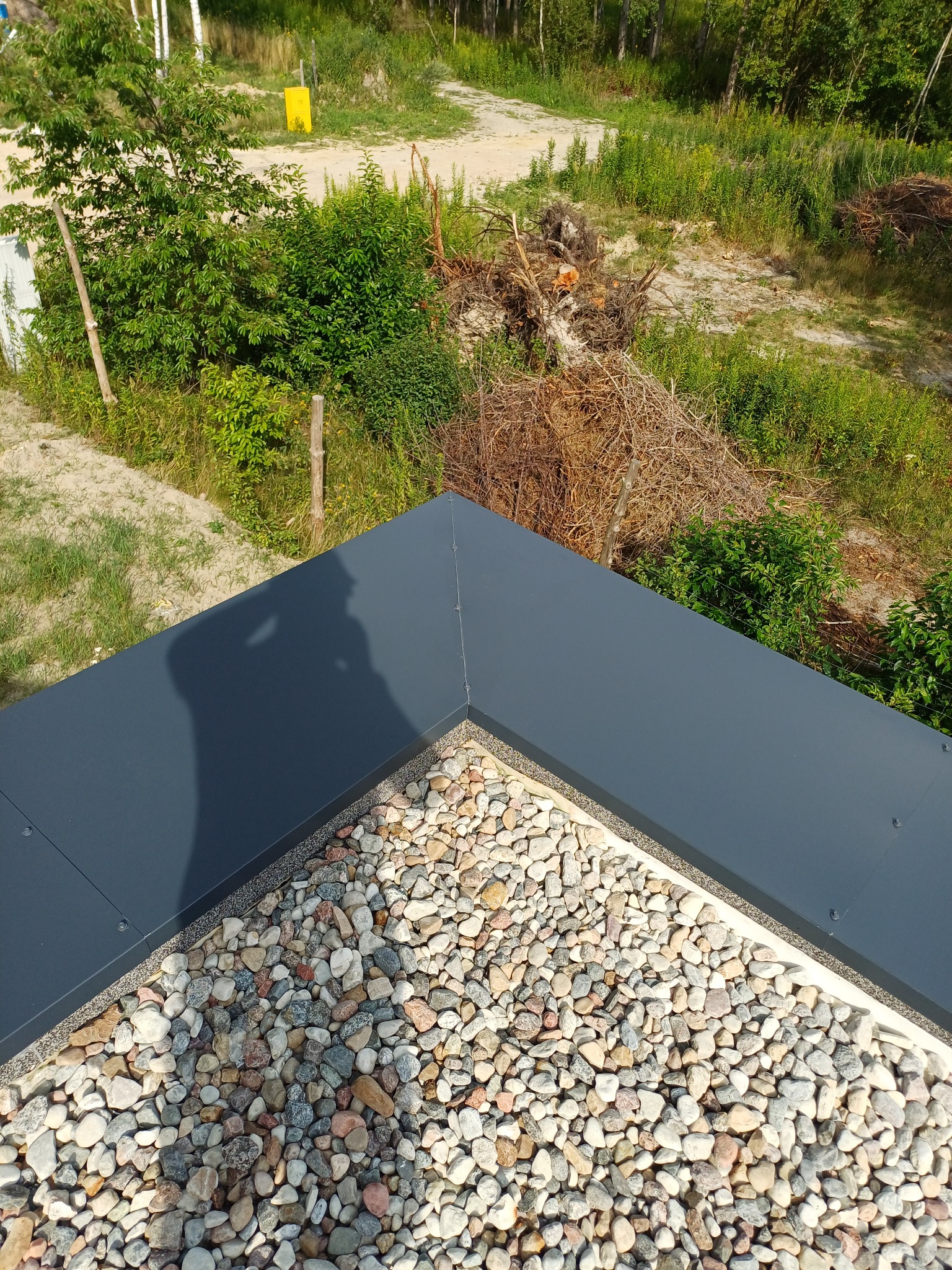 Szczegółowe ujęcie dachu pokrytego drobnymi kamieniami, z ciemną, metalową obróbką blacharską w rogu, cień osoby na dachu i zieleń w tle.