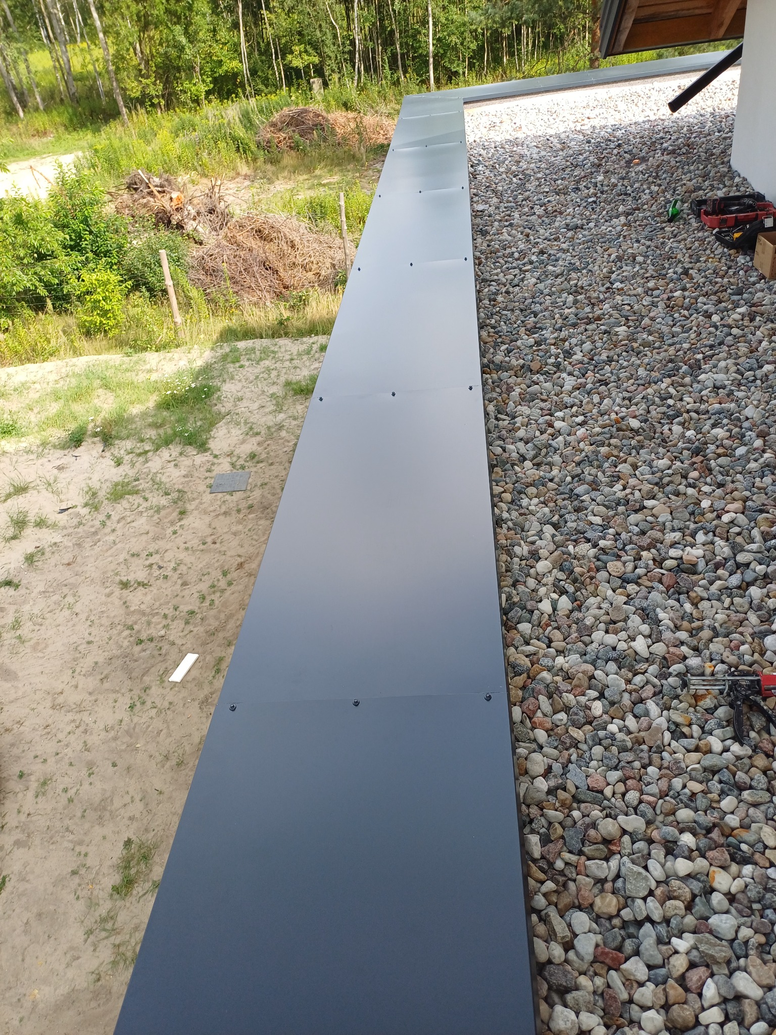 Szara obróbka blacharska na dachu pokrytym kamieniami, widok z góry. Na dachu widoczne narzędzia dekarskie. W tle zieleń i drzewa.