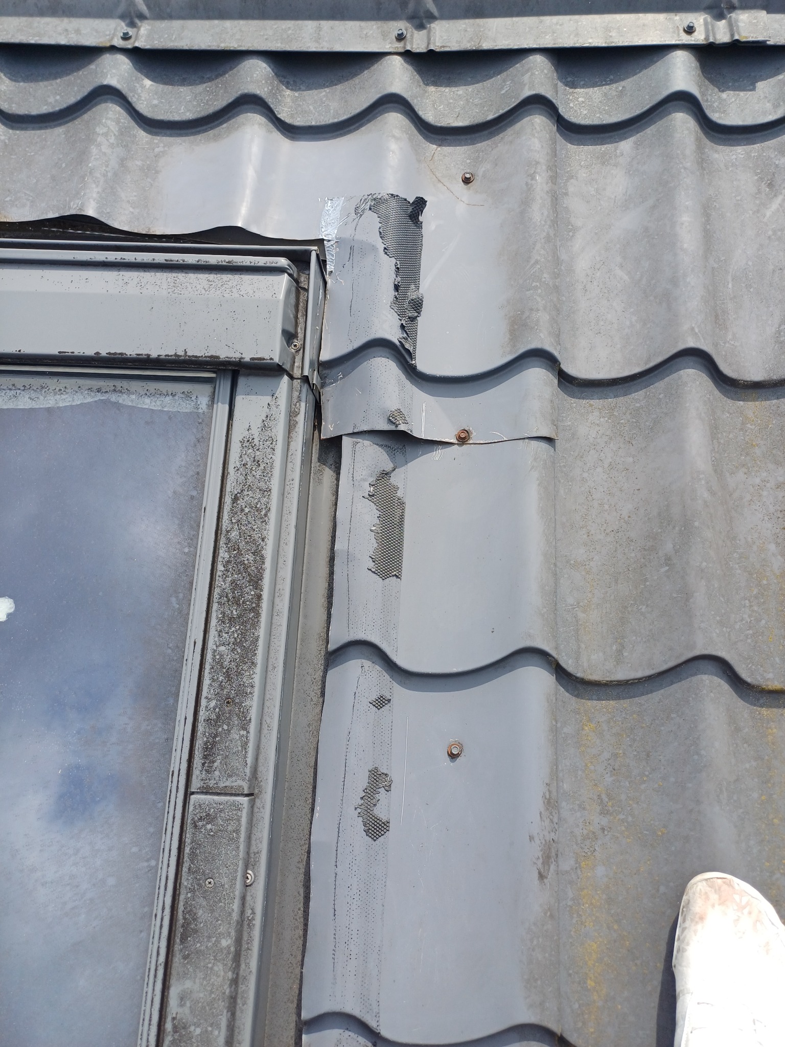 Uszkodzona obróbka blacharska przy oknie dachowym na szarym dachu z dachówki. Widoczne ślady korozji i prowizoryczne naprawy taśmą. Detal uszkodzeń.