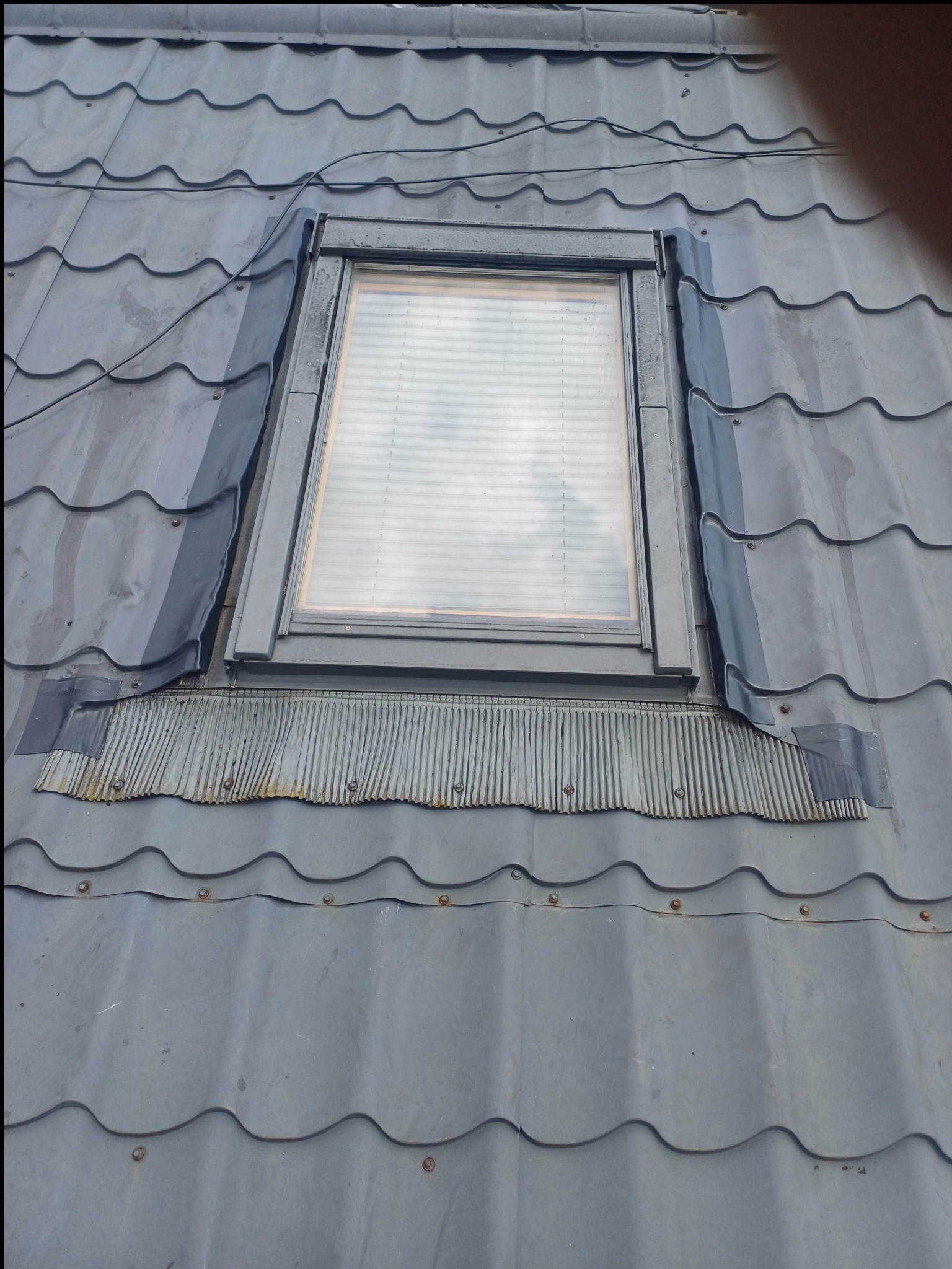 Szare dachówki wokół okna dachowego z widoczną obróbką blacharską i uszczelnieniami, detale mocowań. Widok z dołu na fragment dachu.