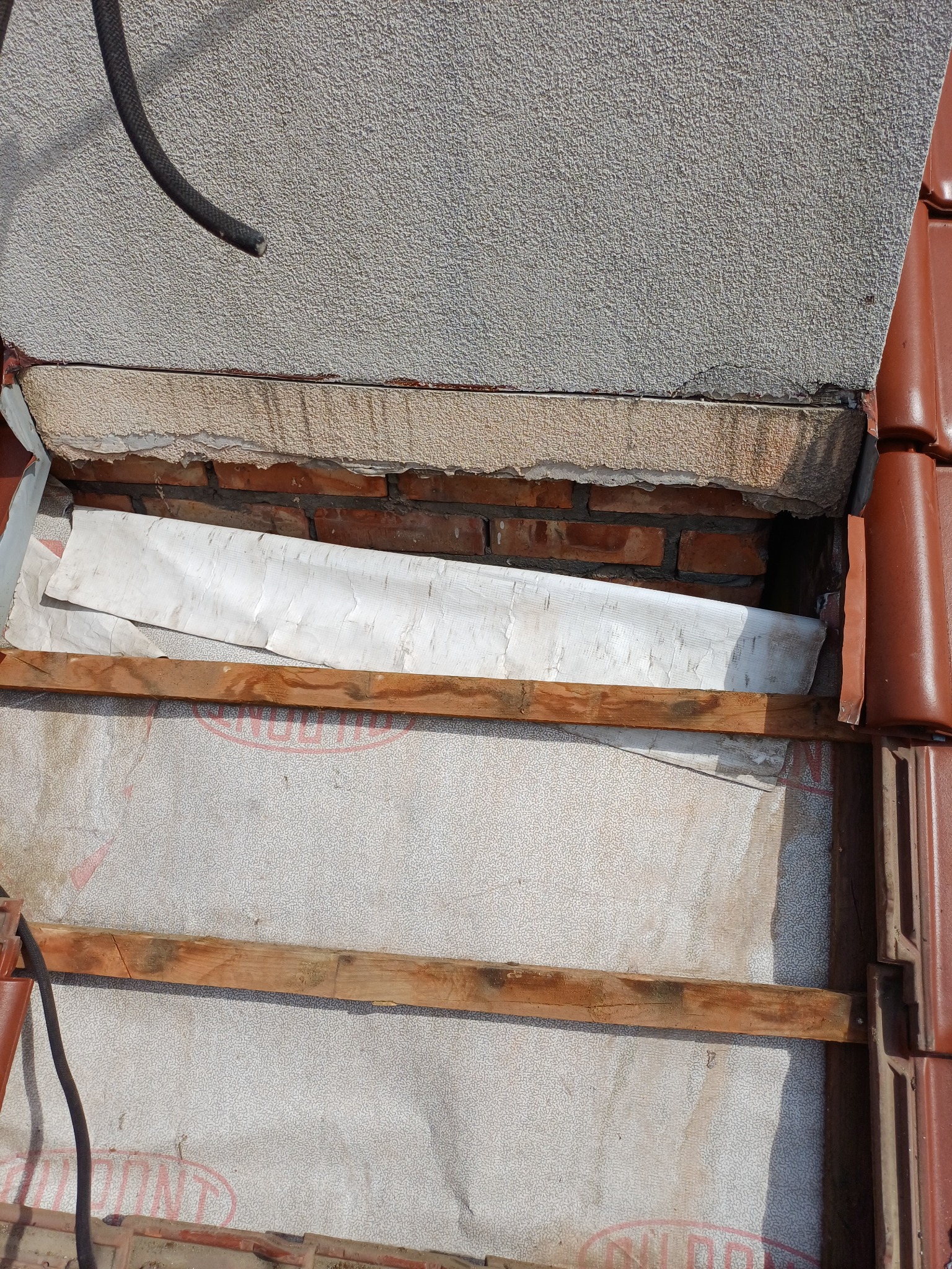 Fragment dachu z widoczną konstrukcją, izolacją i częścią komina. Czerwona dachówka kontrastuje z szarą elewacją. Widoczne drewniane elementy konstrukcyjne.