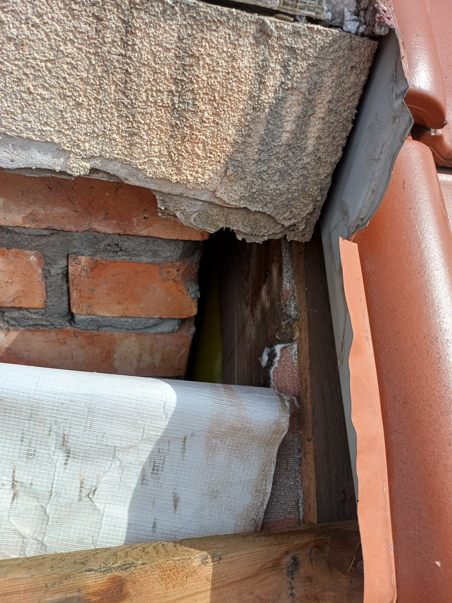 Szczegółowe ujęcie uszkodzonej ściany pod dachem z widoczną cegłą, izolacją i elementami konstrukcji drewnianej. Widoczne ślady zawilgocenia.