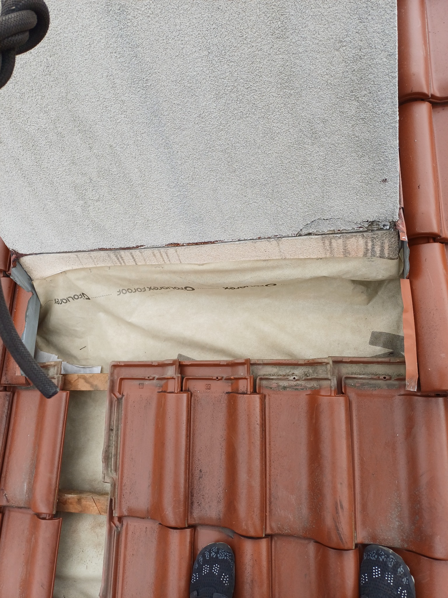 Częściowy widok dachu z czerwoną dachówką, membraną dachową i fragmentem ściany. Widoczne elementy konstrukcyjne i buty osoby fotografującej.