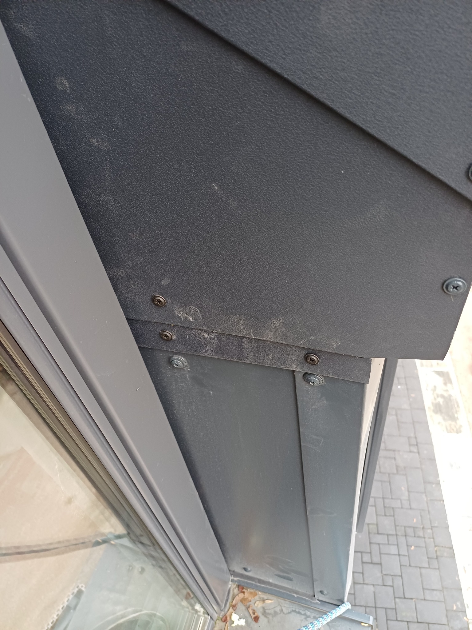 Szczegółowe ujęcie szarej, metalowej obróbki blacharskiej dachu, mocowanej śrubami, z widocznym fragmentem okna i chodnika w tle. Kątowe ujęcie z dołu.