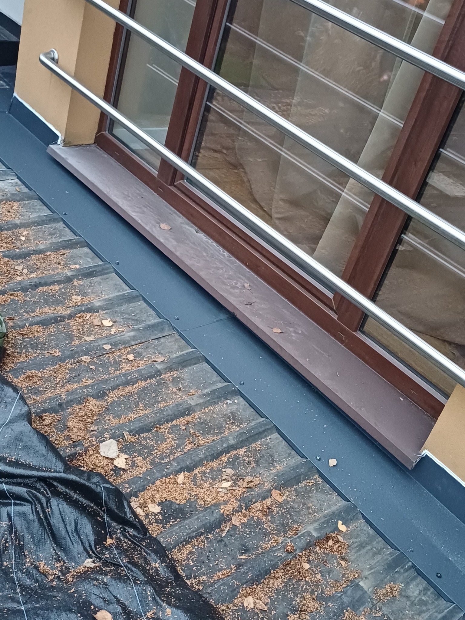 Fragment dachu z oknem połaciowym, zabezpieczony czarną folią. Widoczne liście i drobne zanieczyszczenia na powierzchni dachu. Metalowa barierka przy oknie.