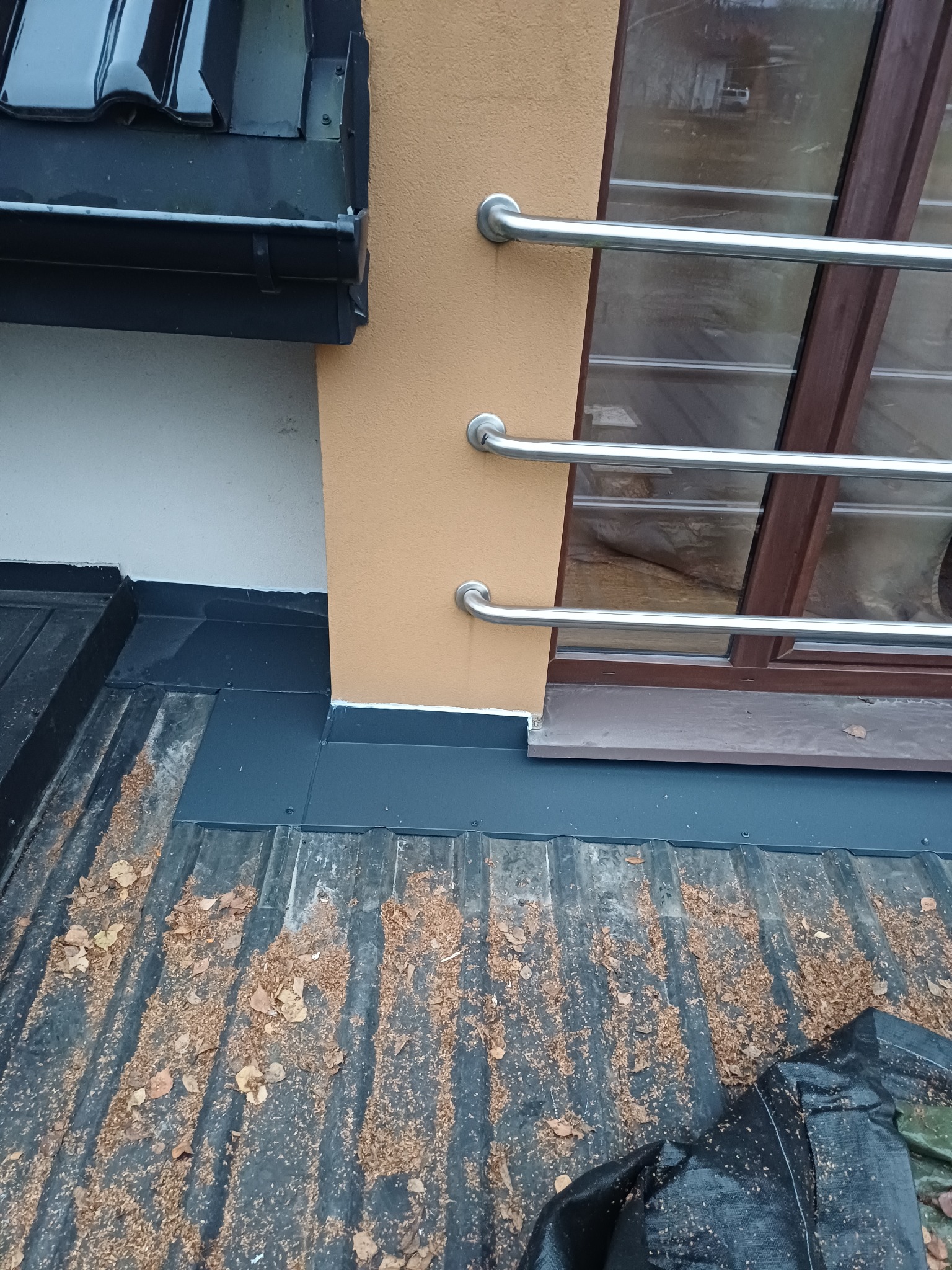 Fragment dachu z blachy trapezowej, z widocznymi liśćmi i fragmentem rynny. W tle okno z metalową barierką i fragment elewacji w ciepłym odcieniu.