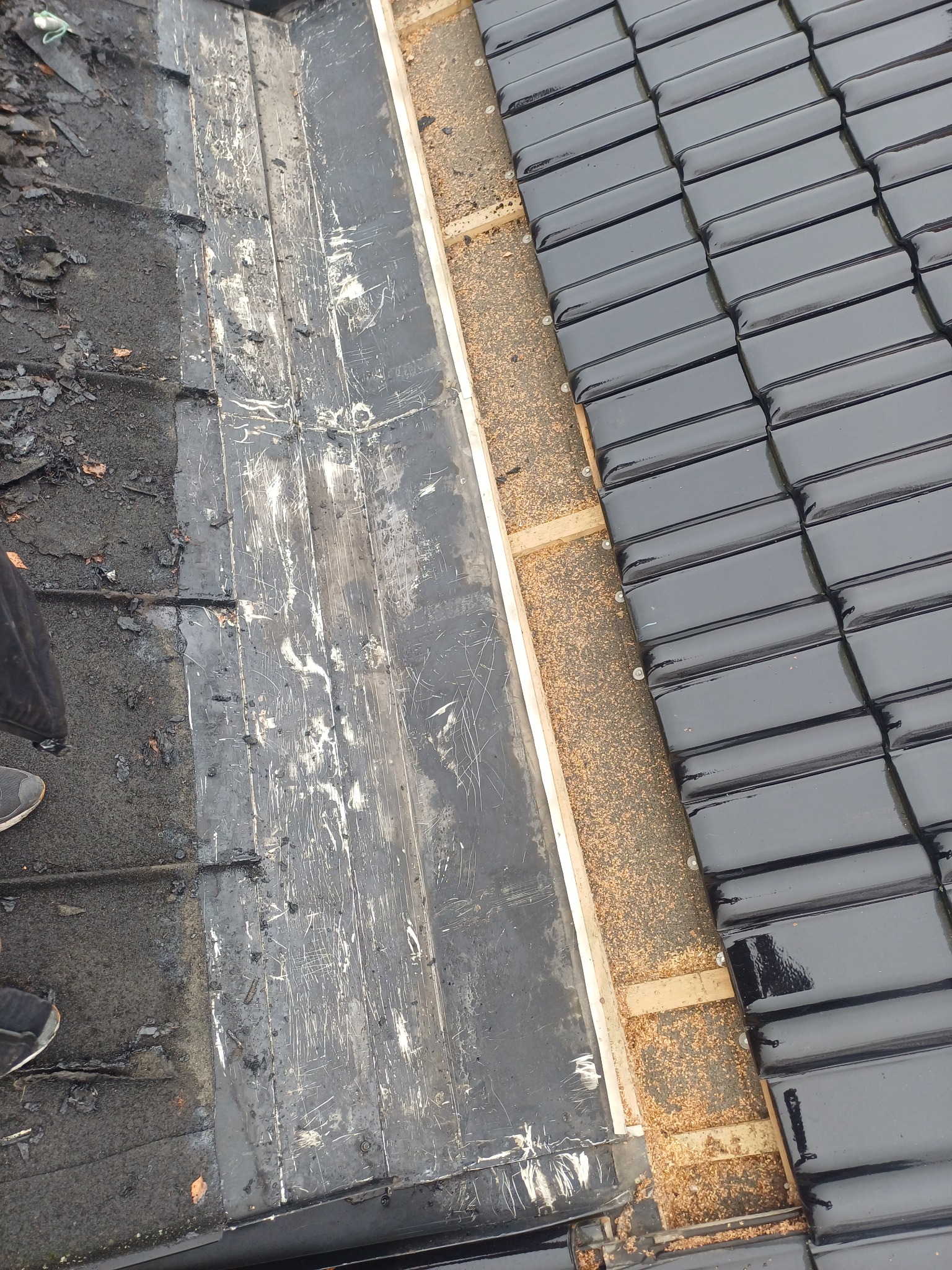 Fragment dachu w trakcie naprawy: stara papa obok nowych, błyszczących dachówek. Widoczne drewniane elementy konstrukcyjne i fragment buta osoby wykonującej prace.