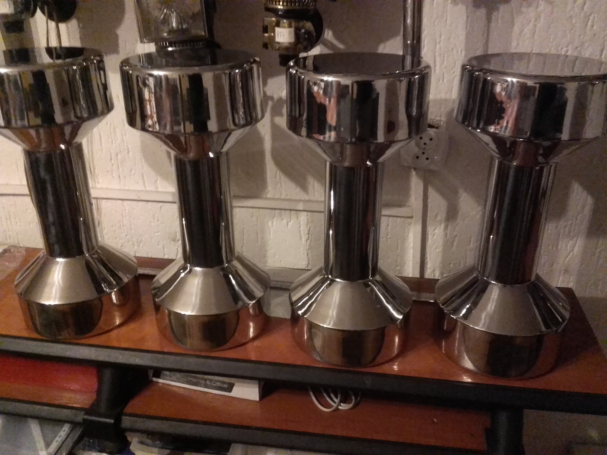 Cztery chromowane, metalowe stojaki o kształcie zbliżonym do hantli, ustawione na drewnianej półce na tle ściany z boazerią. Widoczne odbicia światła.