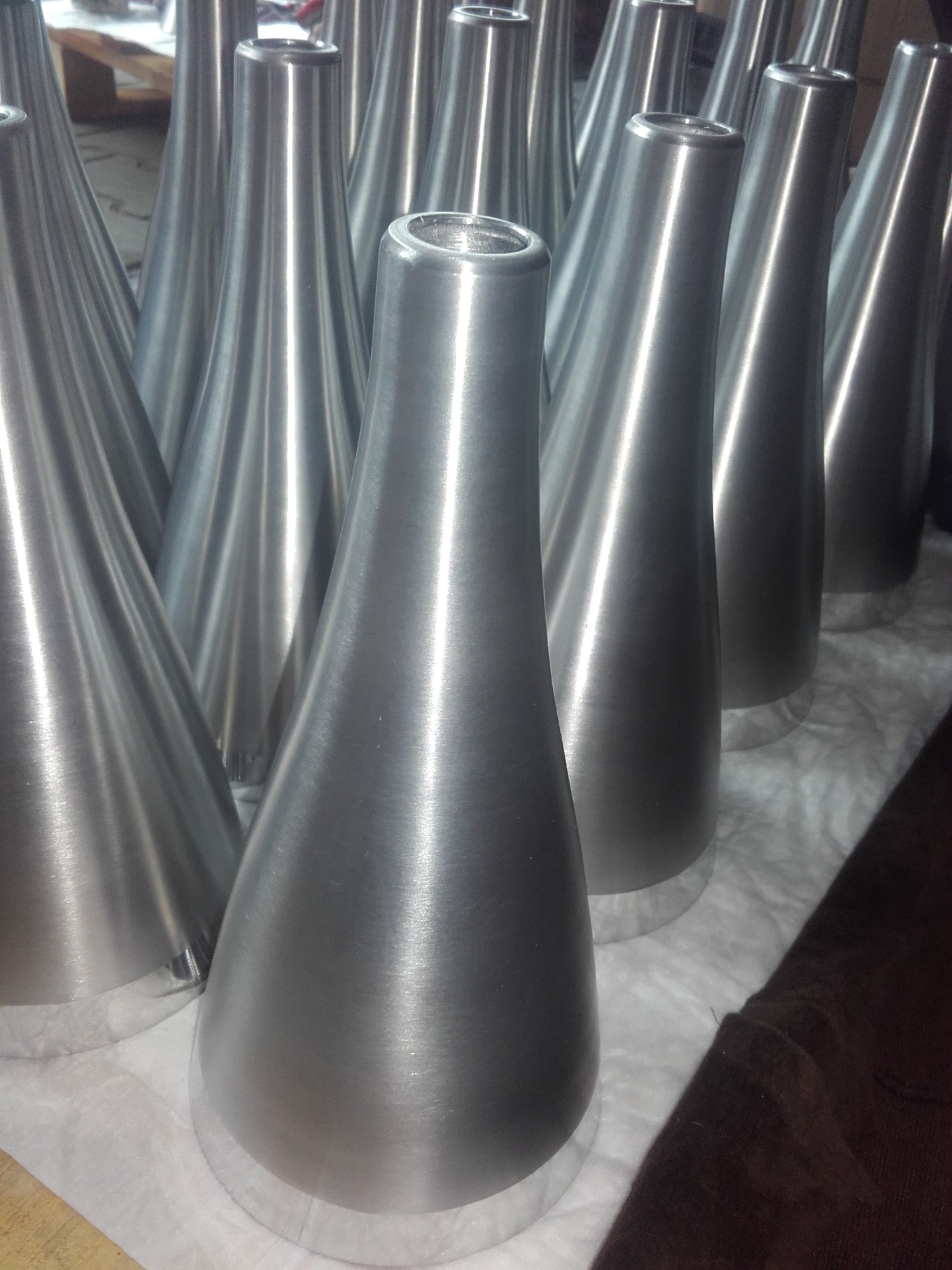 Metalowe, stożkowe elementy z otworem, ustawione rzędem na białym podłożu. Widoczne ślady obróbki mechanicznej na powierzchni.