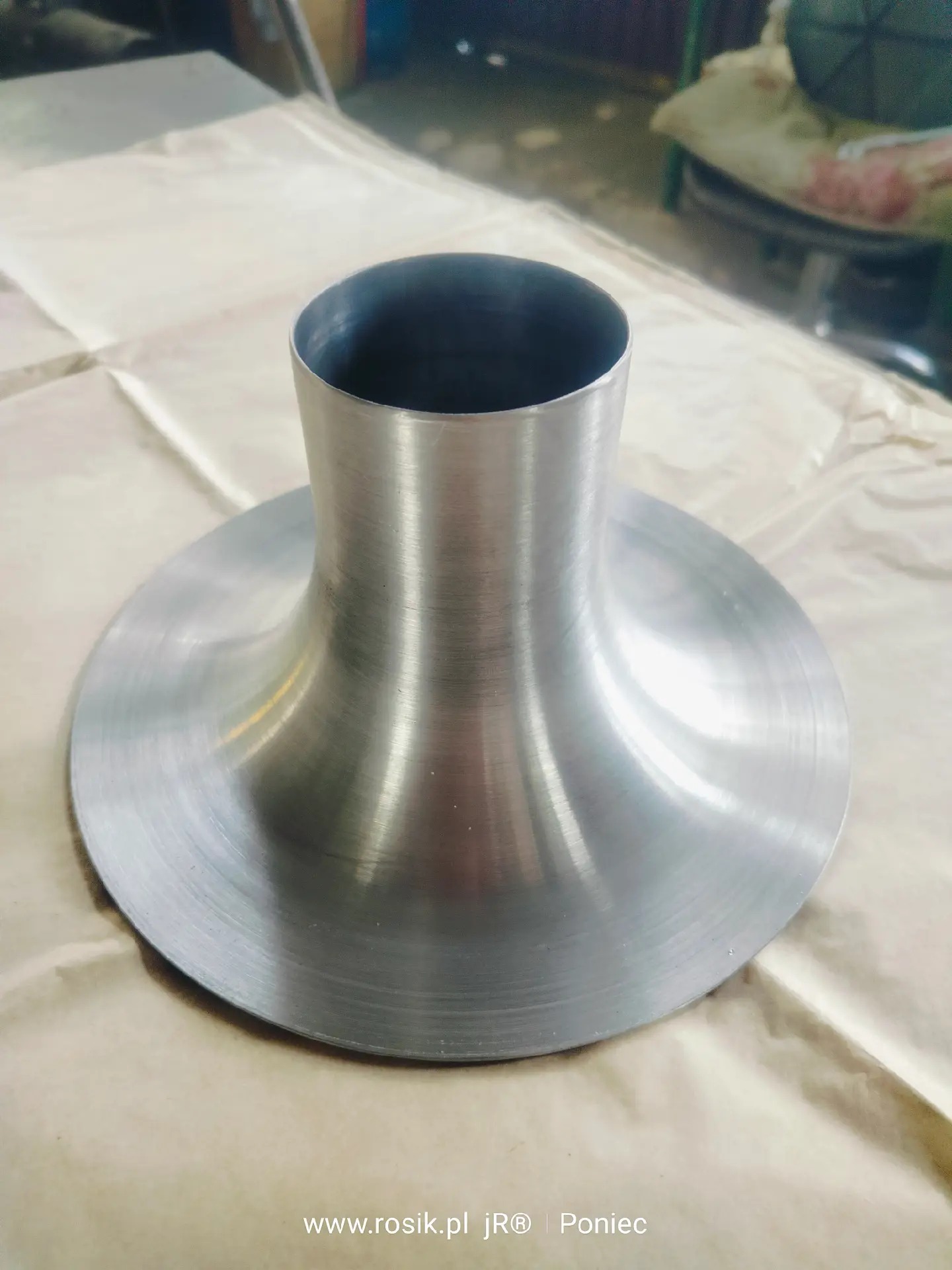 Metalowy element o kształcie stożka z kołnierzem, wykonany z polerowanego metalu. Widoczne ślady obróbki na powierzchni.  Zdjęcie z bliska, na neutralnym tle.