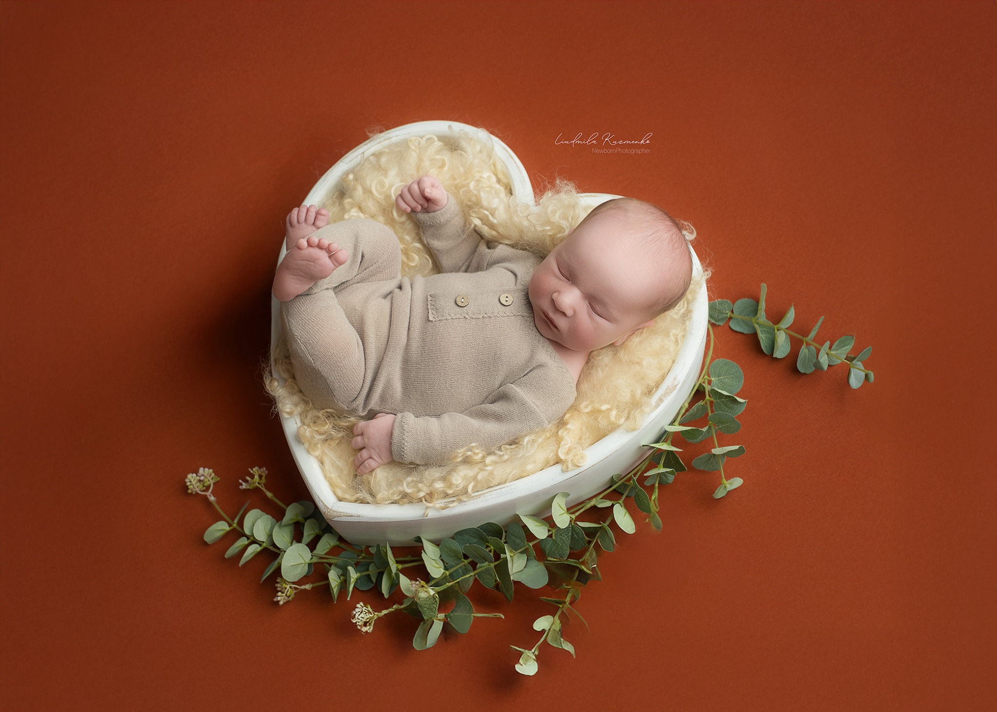 Śpiący noworodek w beżowym ubranku leży w białym sercu z kremowym futerkiem, otoczonym zielonymi gałązkami na pomarańczowym tle.