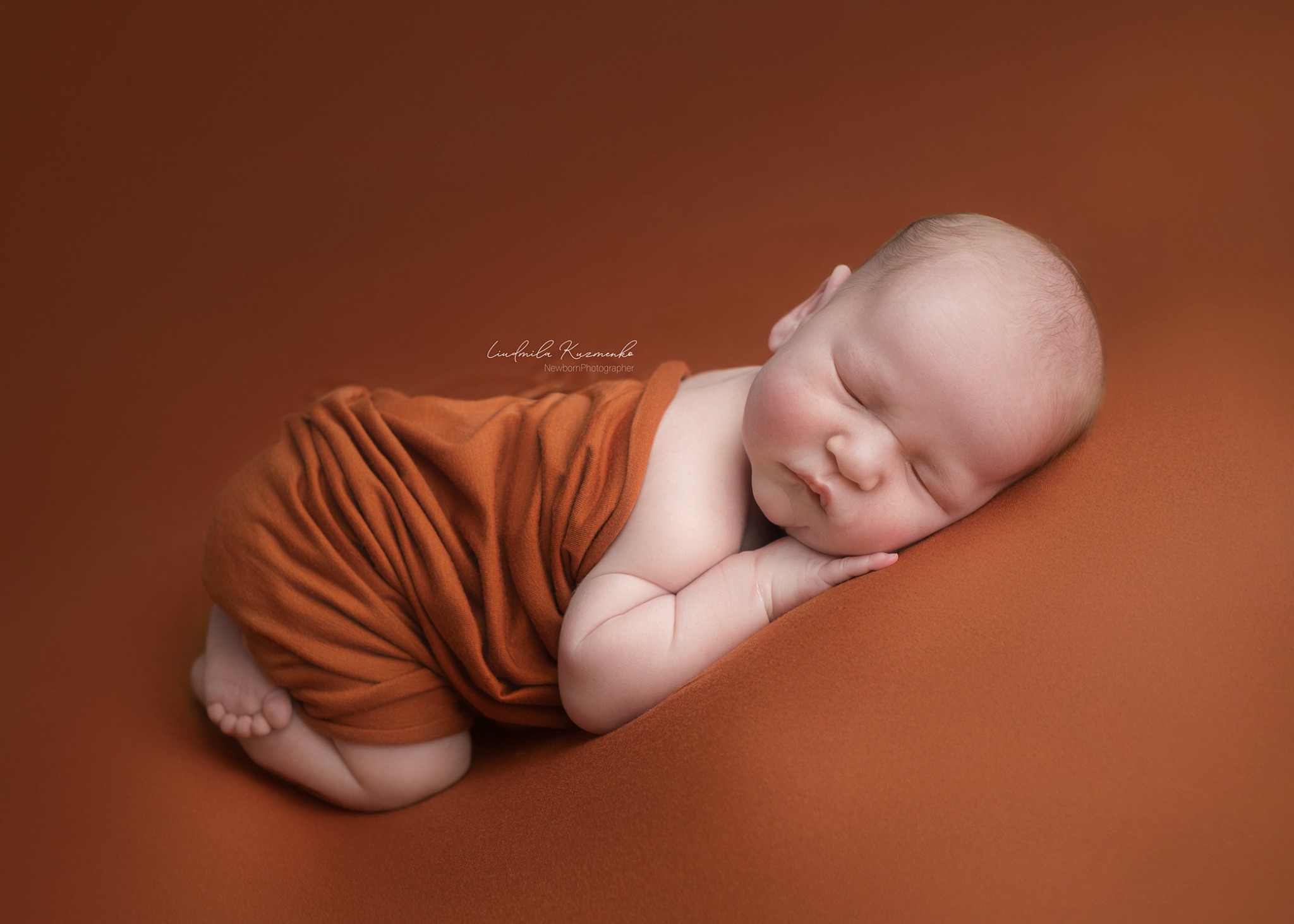 Śpiący noworodek na brązowym tle, owinięty w pomarańczową tkaninę. Dziecko leży na boku z rączką pod policzkiem. Delikatne oświetlenie podkreśla naturalne rysy twarzy.