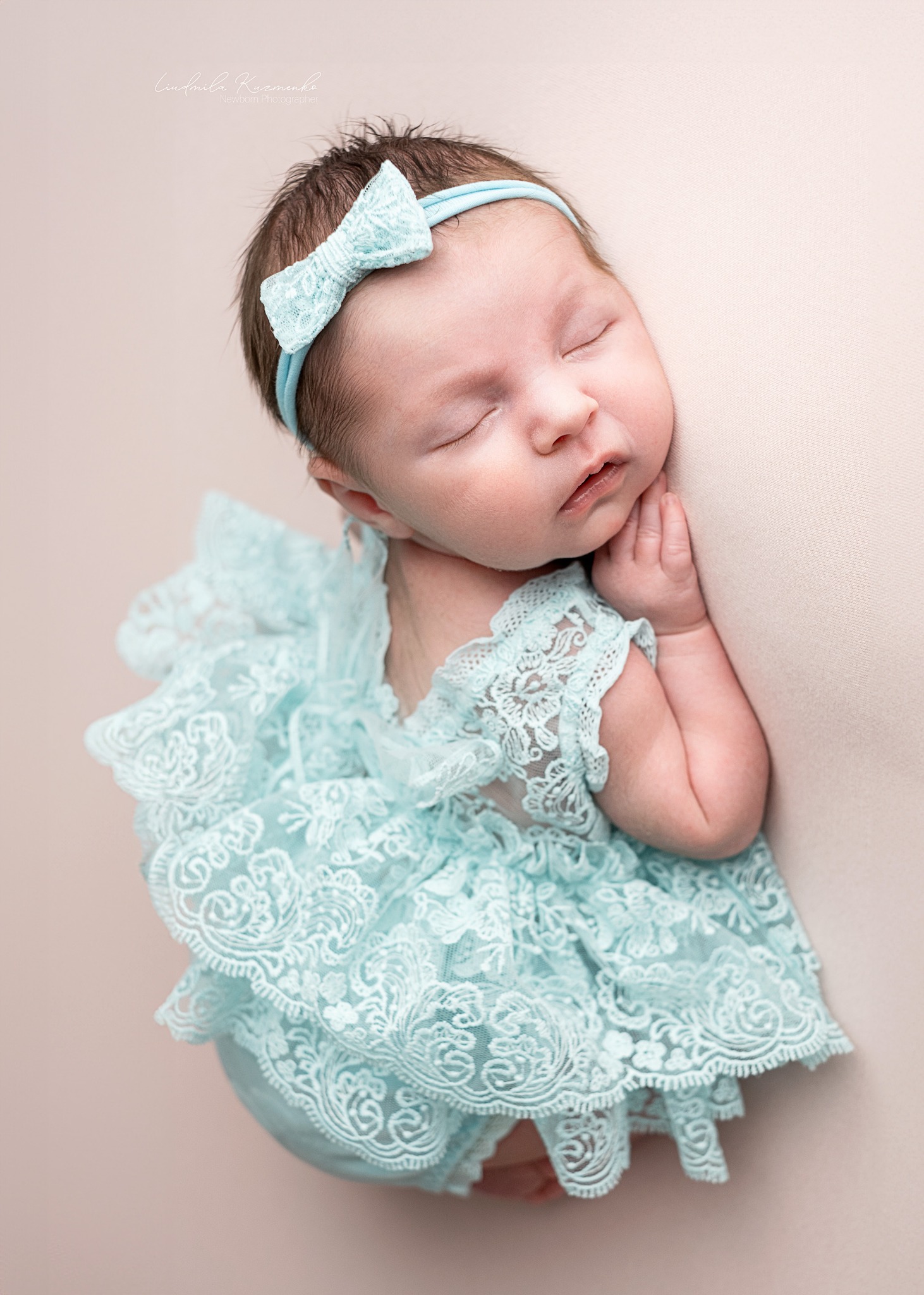 Śpiący noworodek w błękitnej koronkowej sukience i opasce z kokardką, ułożony na beżowym tle. Dziecko ma złożone rączki przy policzku. Delikatne oświetlenie podkreśla detale.