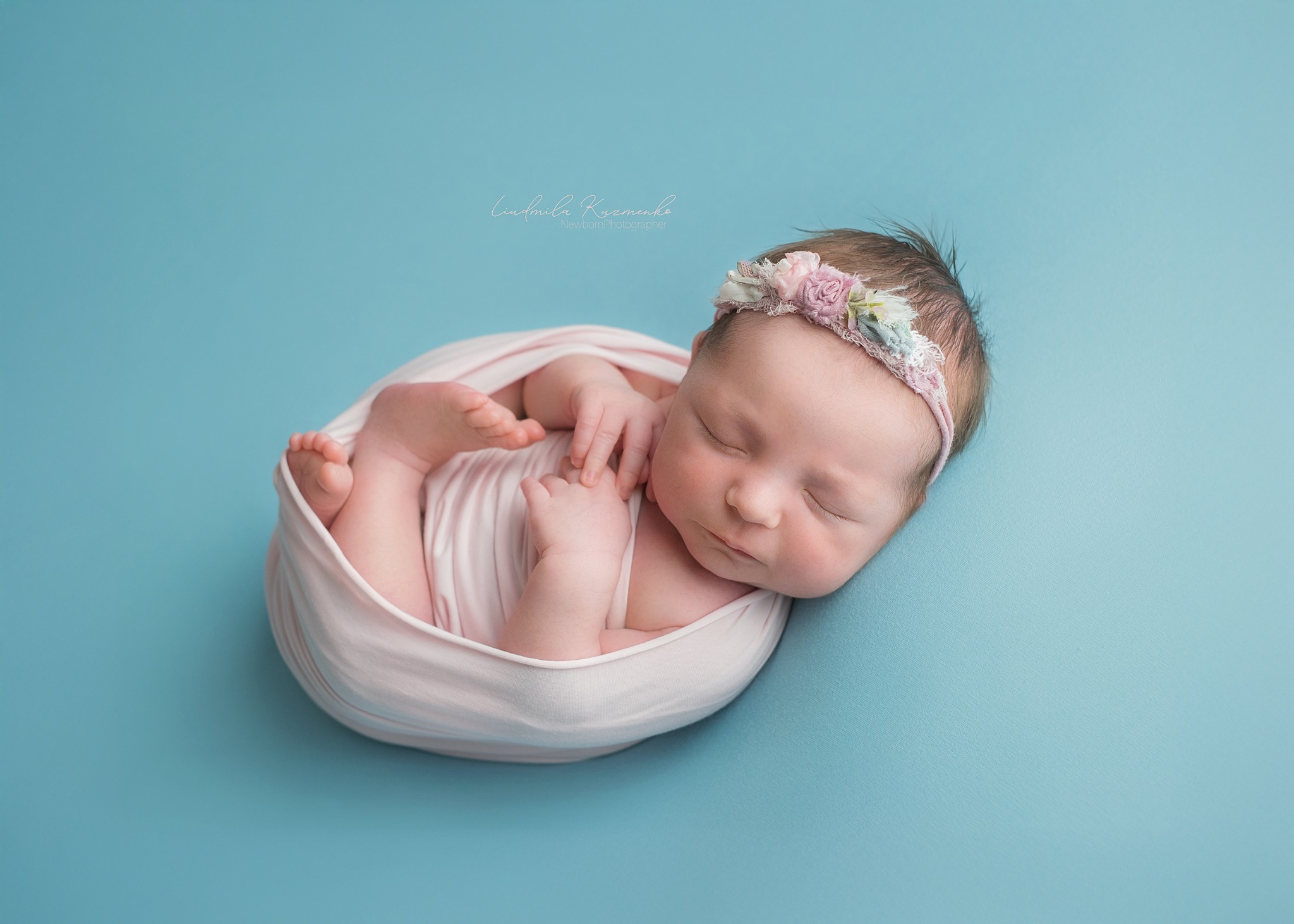 Śpiący noworodek w kokonie z różowej tkaniny, ozdobiony kwiatową opaską na głowie, na gładkim, błękitnym tle. Delikatne światło podkreśla rysy twarzy.