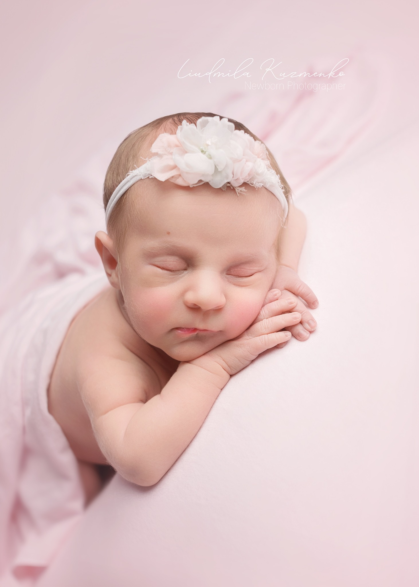 Śpiąca noworodka z kwiatową opaską na głowie, owinięta w delikatny różowy materiał, leżąca na miękkiej poduszce w pastelowym odcieniu. Delikatne światło podkreśla jej rysy.