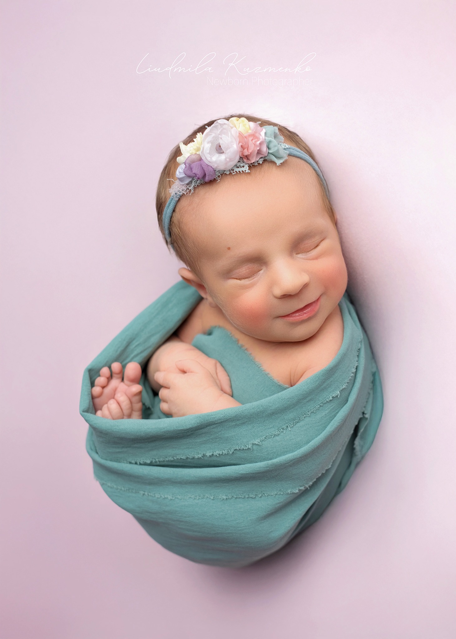 Uśmiechnięty noworodek w turkusowym otulaczu i z kwiatową opaską na głowie, na delikatnym różowym tle. Zdjęcie ze Stargardu.
