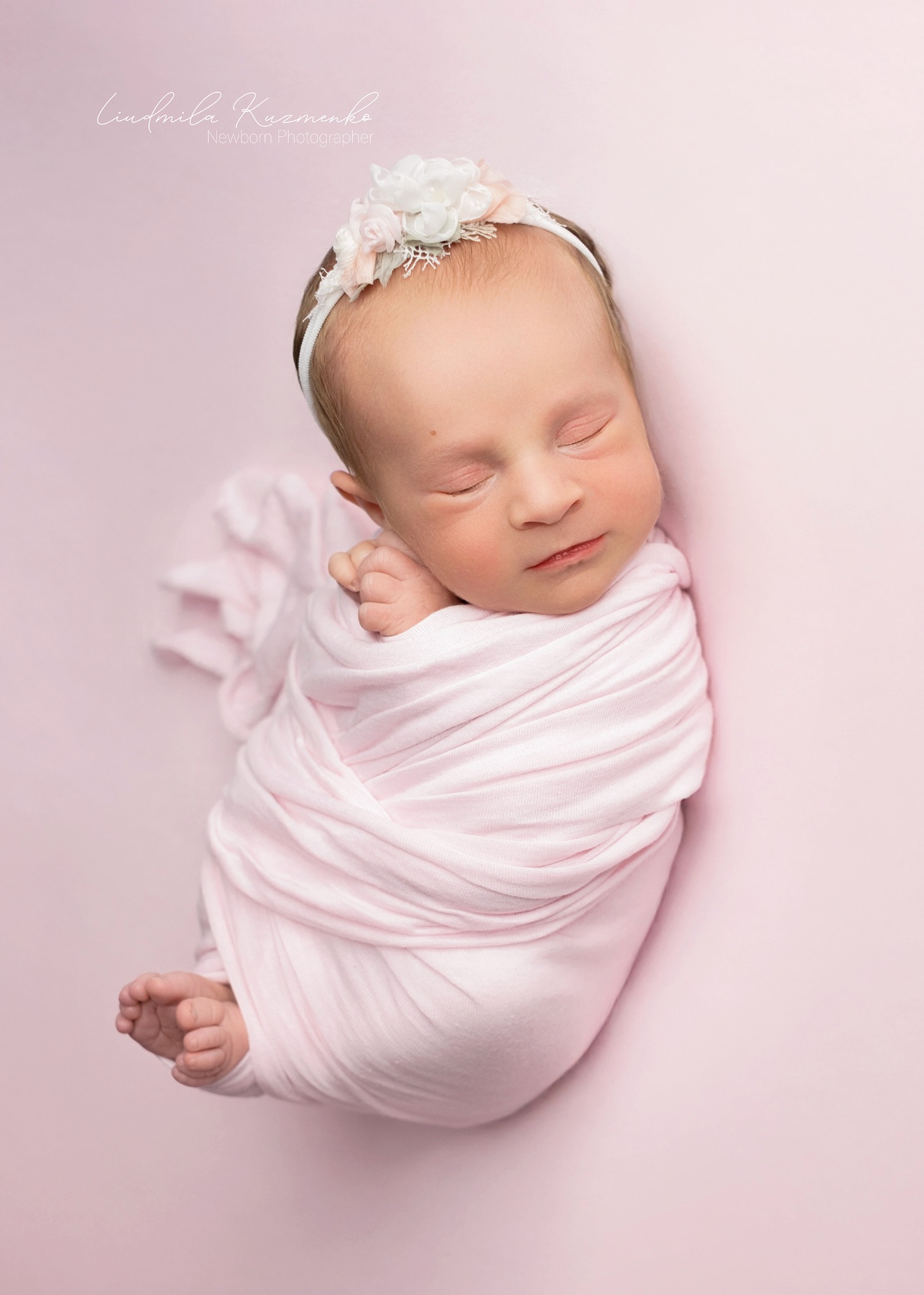 Śpiący noworodek zawinięty w różowy materiał, z opaską w kwiaty na głowie, sfotografowany na jednolitym, jasnoróżowym tle. Delikatne światło podkreśla rysy twarzy.