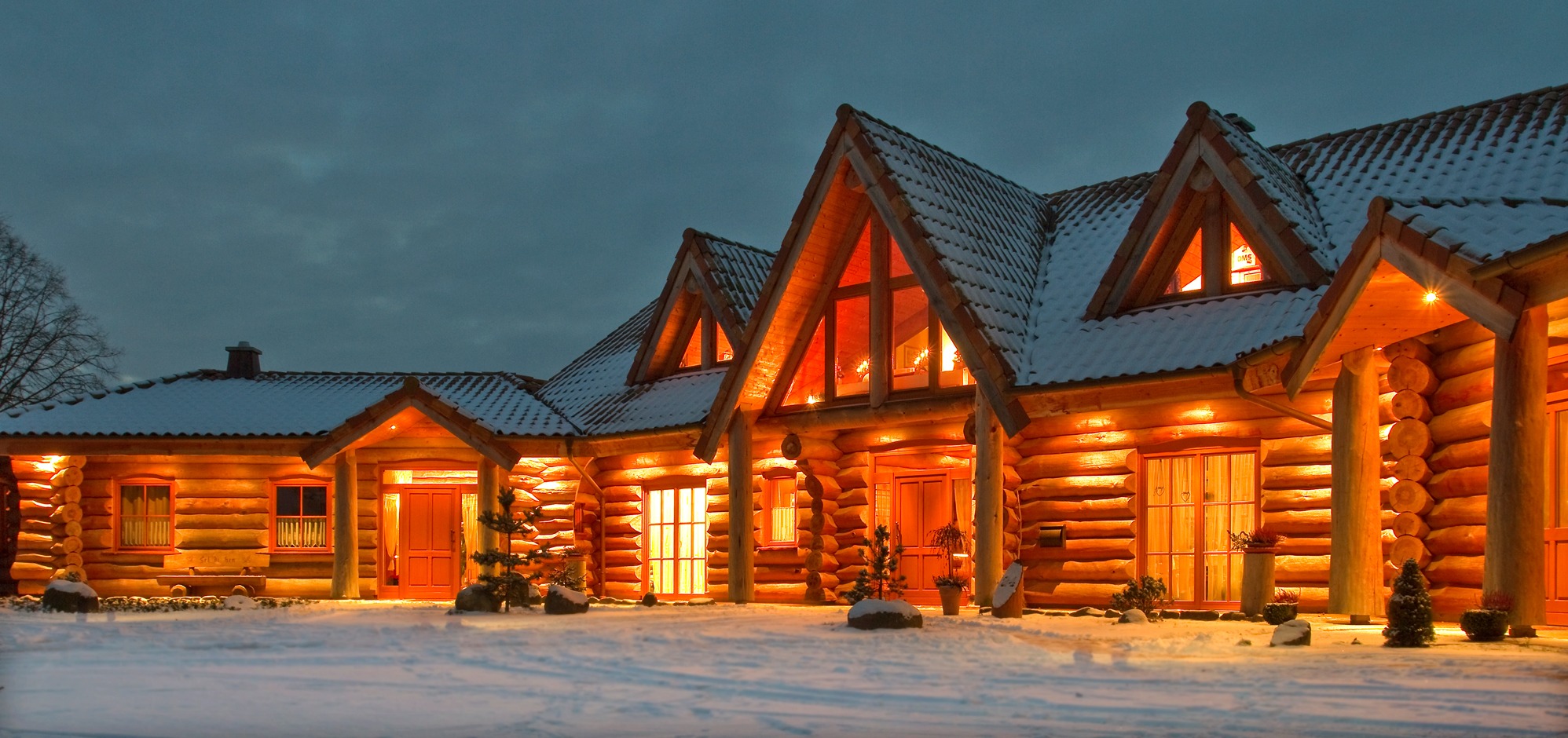 Zimowy krajobraz: Oświetlony dom z bali z dachem pokrytym śniegiem, widoczny o zmierzchu. Ciepłe światło w oknach kontrastuje z chłodem otoczenia.