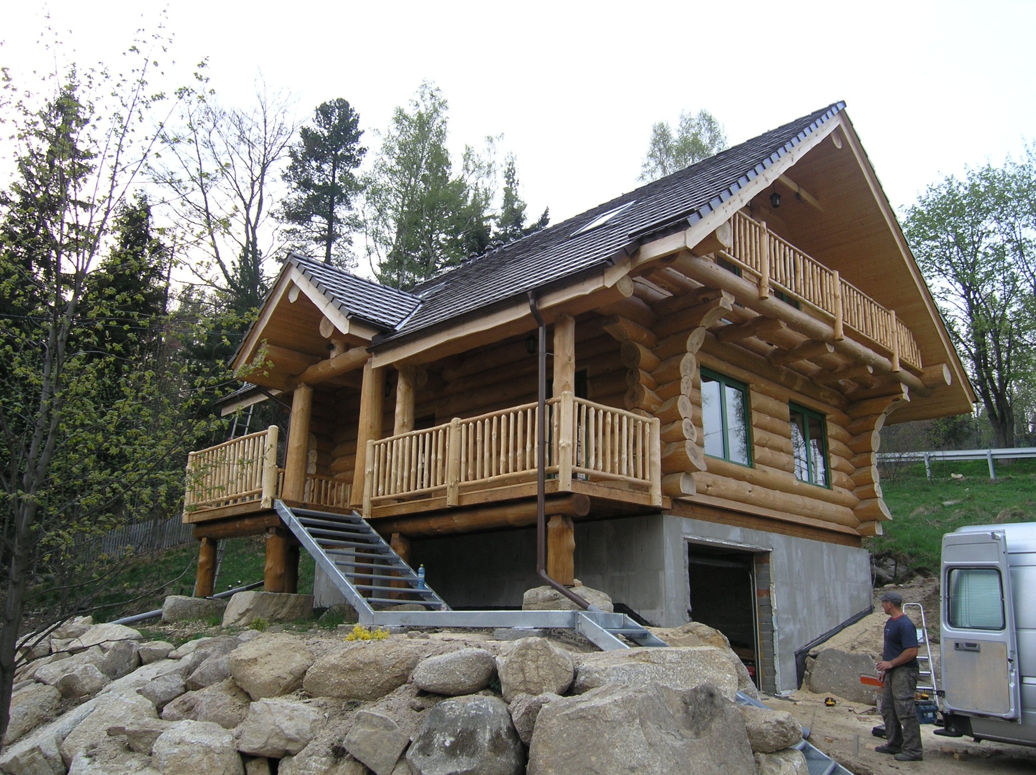 Dwukondygnacyjny dom z bali z balkonami, osadzony na kamiennym fundamencie, z metalowymi schodami i pracownikiem obok srebrnego vana.