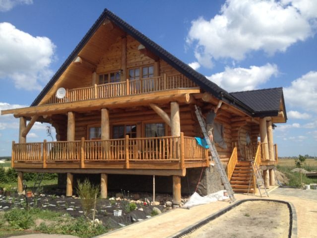 Nowoczesny dom z bali z czarnym dachem, balkonem i kamienną podmurówką, w trakcie prac wykończeniowych przy użyciu drabiny i narzędzi.