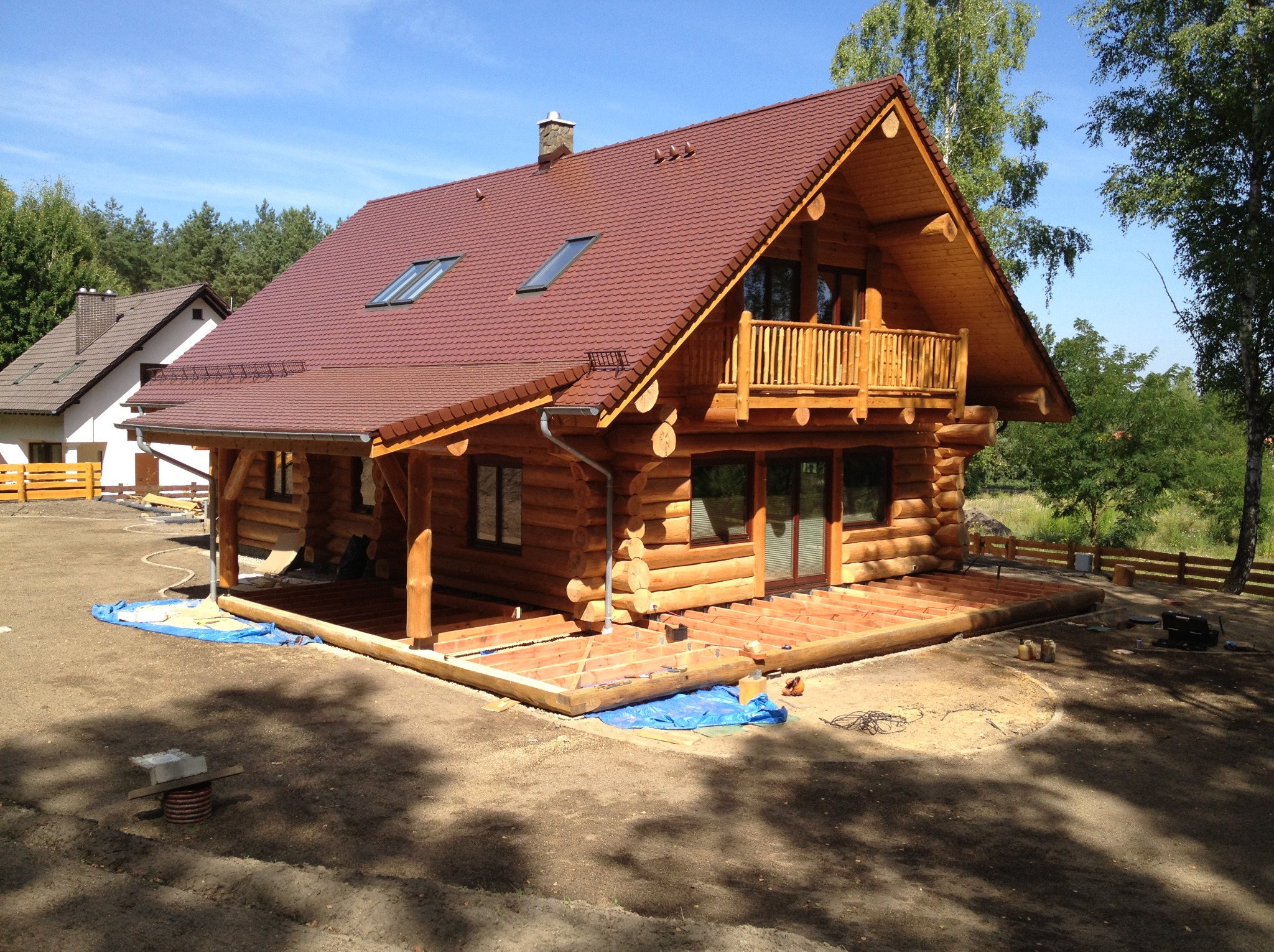 Drewniany dom z bali z balkonem i brązowym dachem w słoneczny dzień. Widoczny fragment konstrukcji tarasu i sąsiedni budynek w tle.