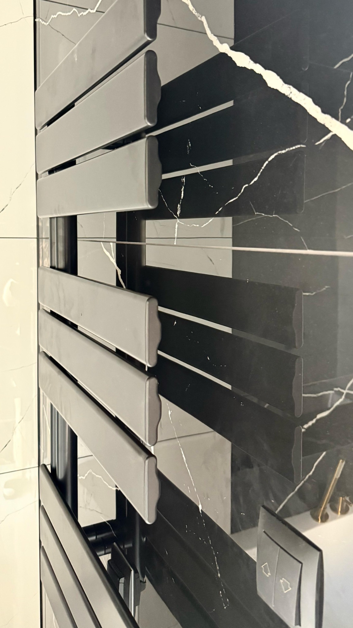 Nowoczesny grzejnik łazienkowy w szarości, na tle ściany wykończonej czarnym marmurem z białymi żyłami. Widoczny fragment białej ściany i włącznik światła.