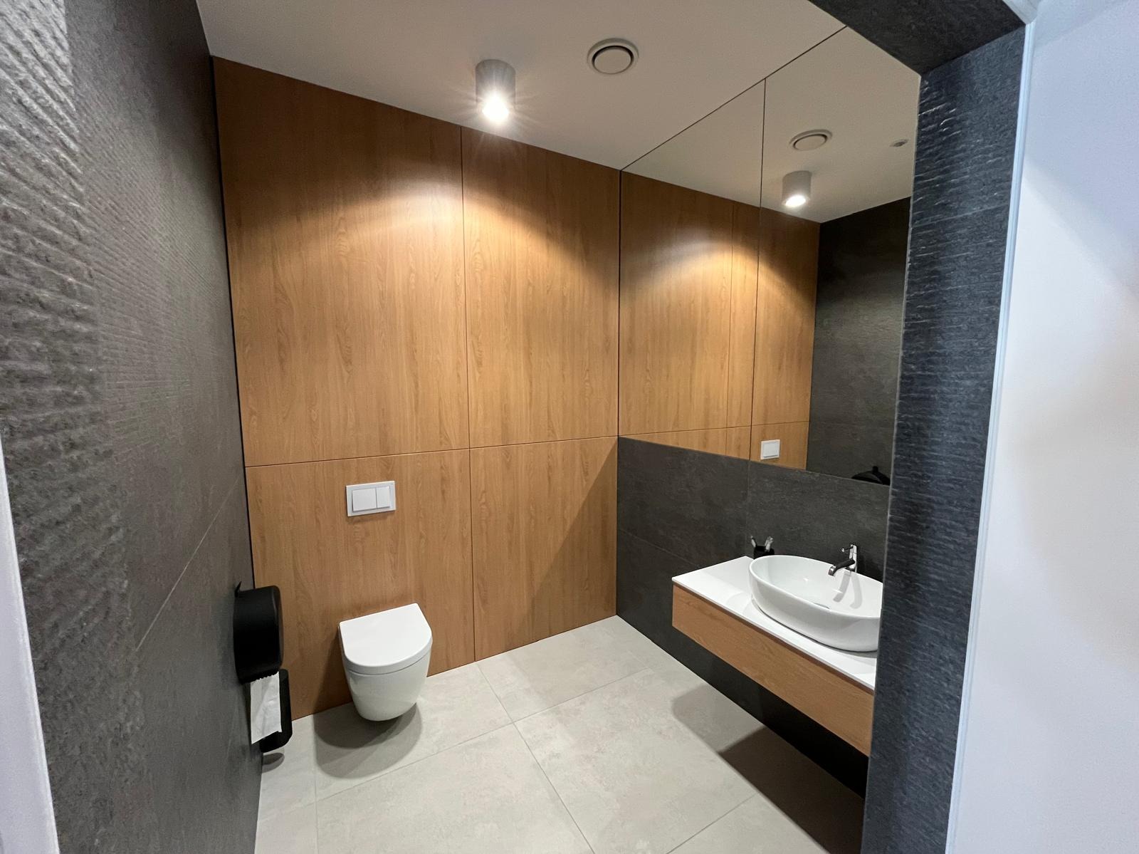 Nowoczesna łazienka z drewnianą okładziną ścienną, szarą płytką imitującą kamień i owalną umywalką nablatową. Minimalistyczny design, ciepłe oświetlenie punktowe.