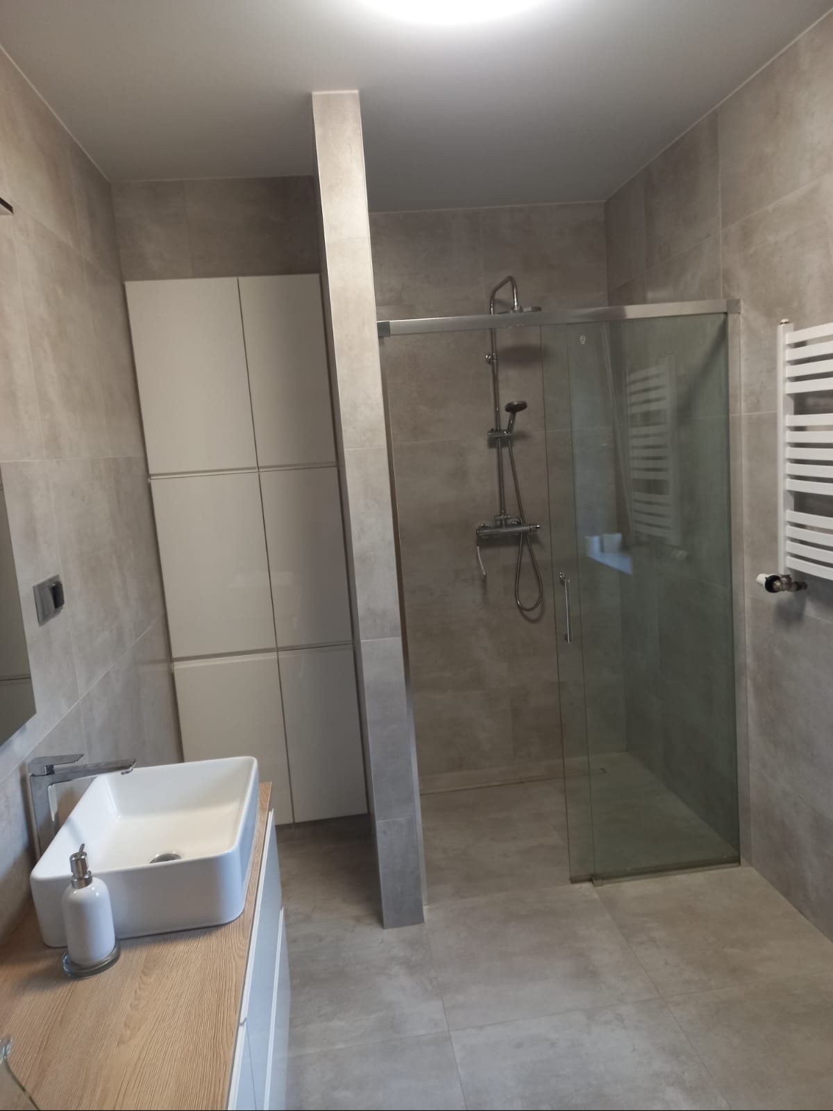 Nowoczesna łazienka w szarościach: kabina prysznicowa ze szklanymi drzwiami, umywalka nablatowa i biała zabudowa meblowa tworzą spójną, minimalistyczną przestrzeń.