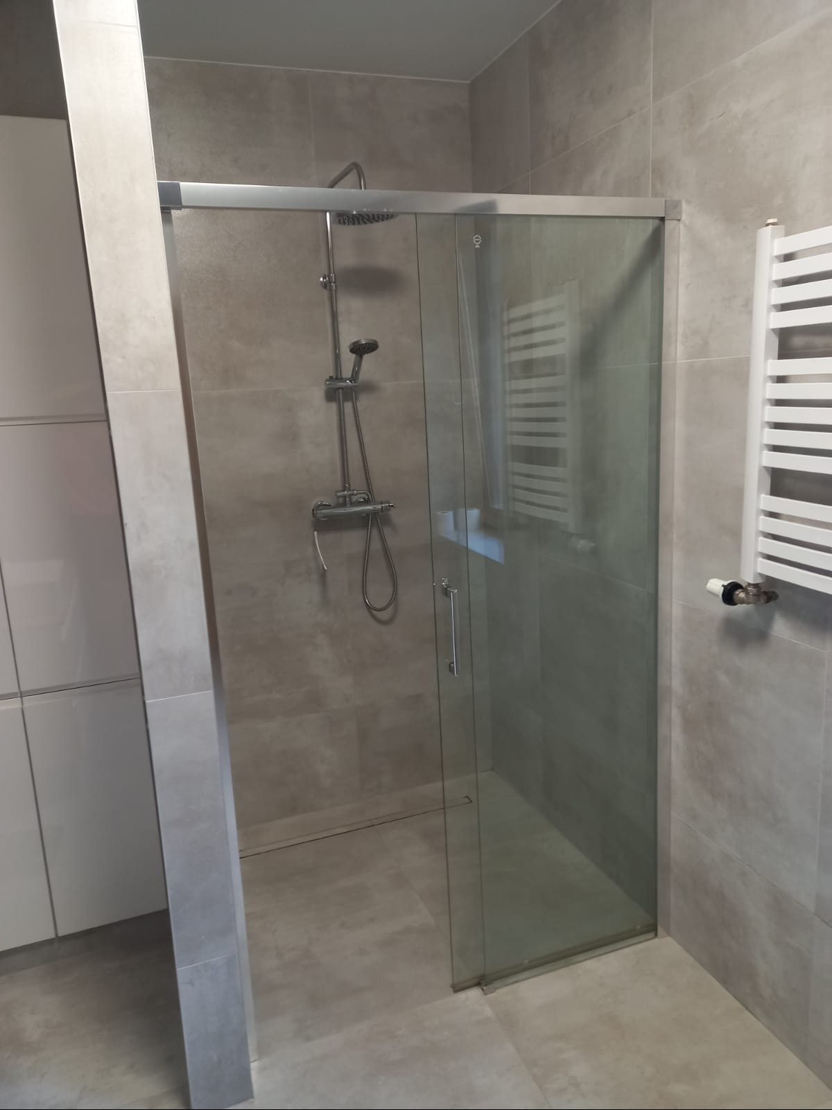Szklana kabina prysznicowa z przesuwnymi drzwiami, srebrne okucia, szara płytka imitująca beton na ścianach i podłodze. Minimalistyczny design, biały grzejnik drabinkowy.
