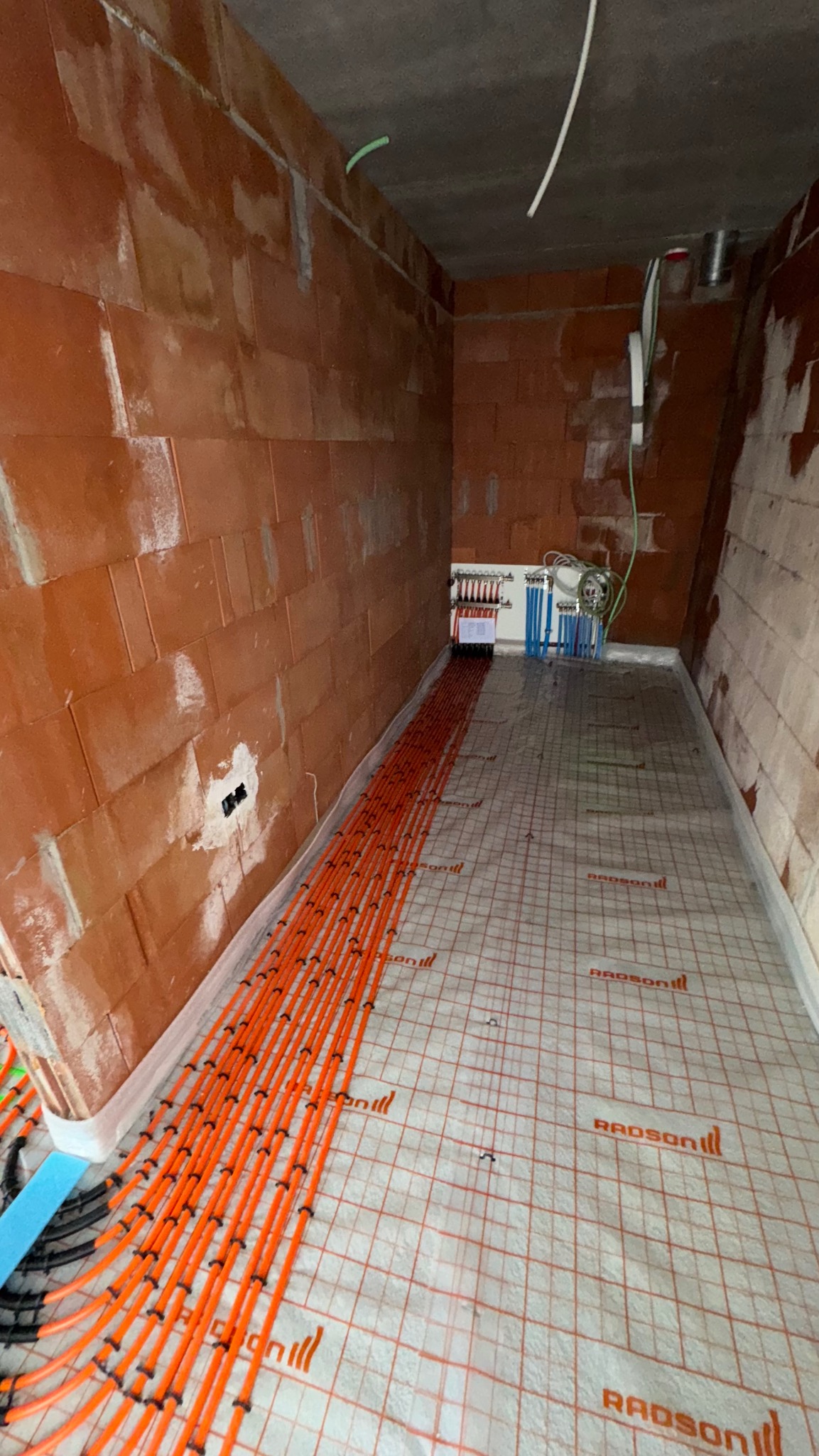 Pomarańczowe rurki ogrzewania podłogowego Radson ułożone na macie izolacyjnej w surowym pomieszczeniu z ceglanymi ścianami. Widoczny rozdzielacz.