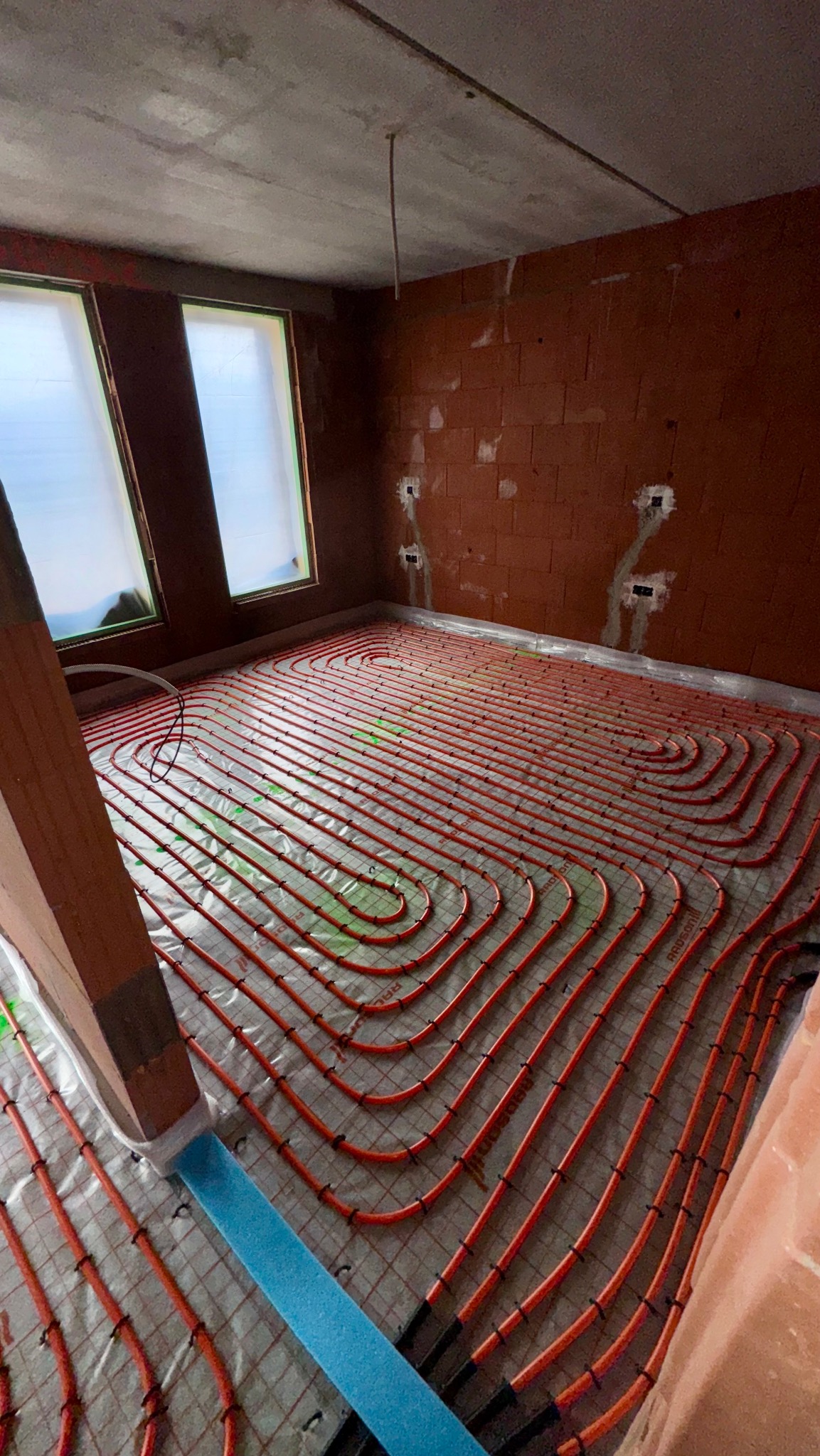 Pomarańczowe rury ogrzewania podłogowego ułożone spiralnie na folii w surowym pomieszczeniu z oknami i ceglanymi ścianami. Widoczny fragment drewnianej konstrukcji.