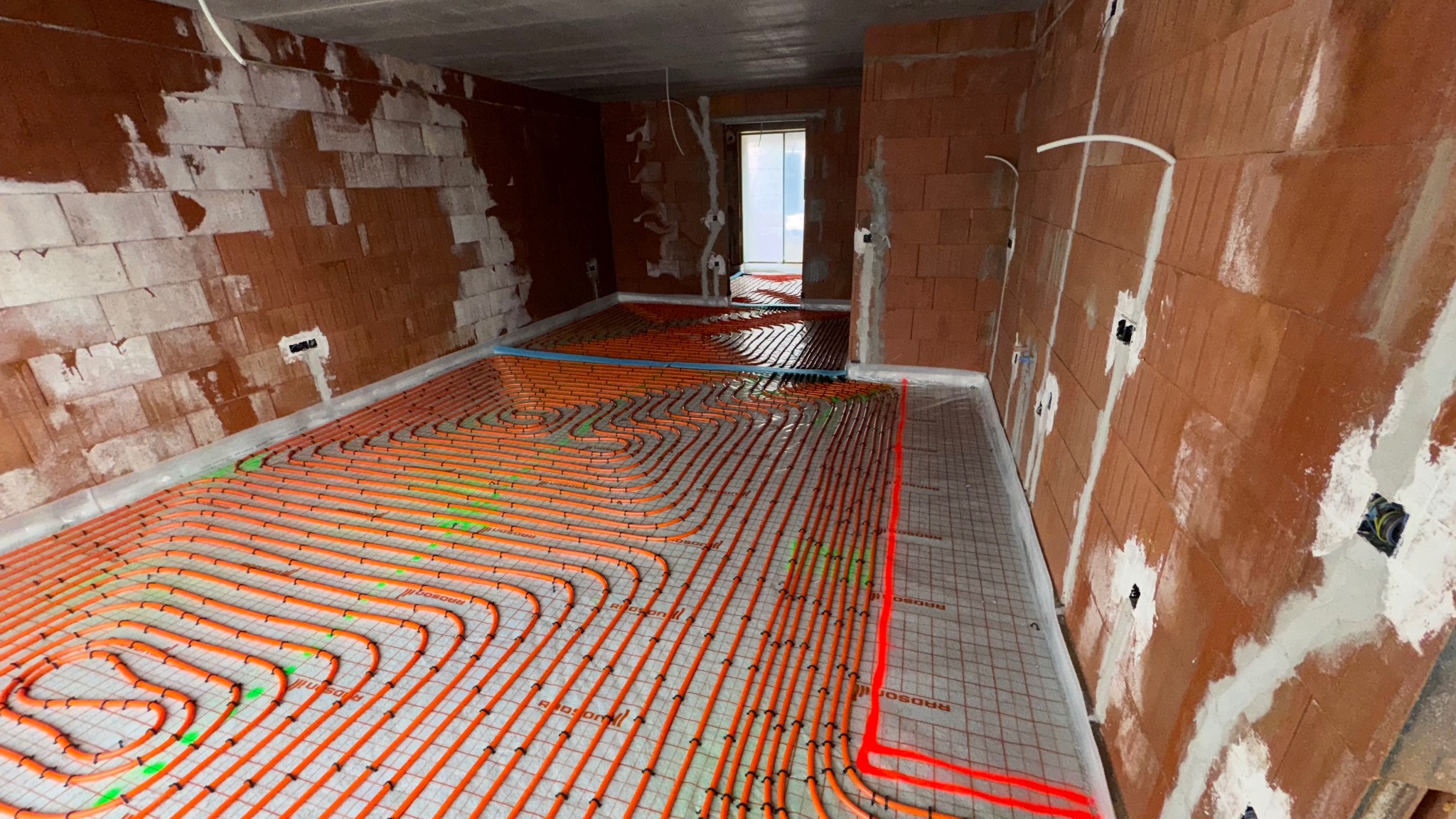 Pomarańczowe rury ogrzewania podłogowego ułożone spiralnie na folii z siatką w surowym wnętrzu z ceglanymi ścianami, widoczne instalacje elektryczne.